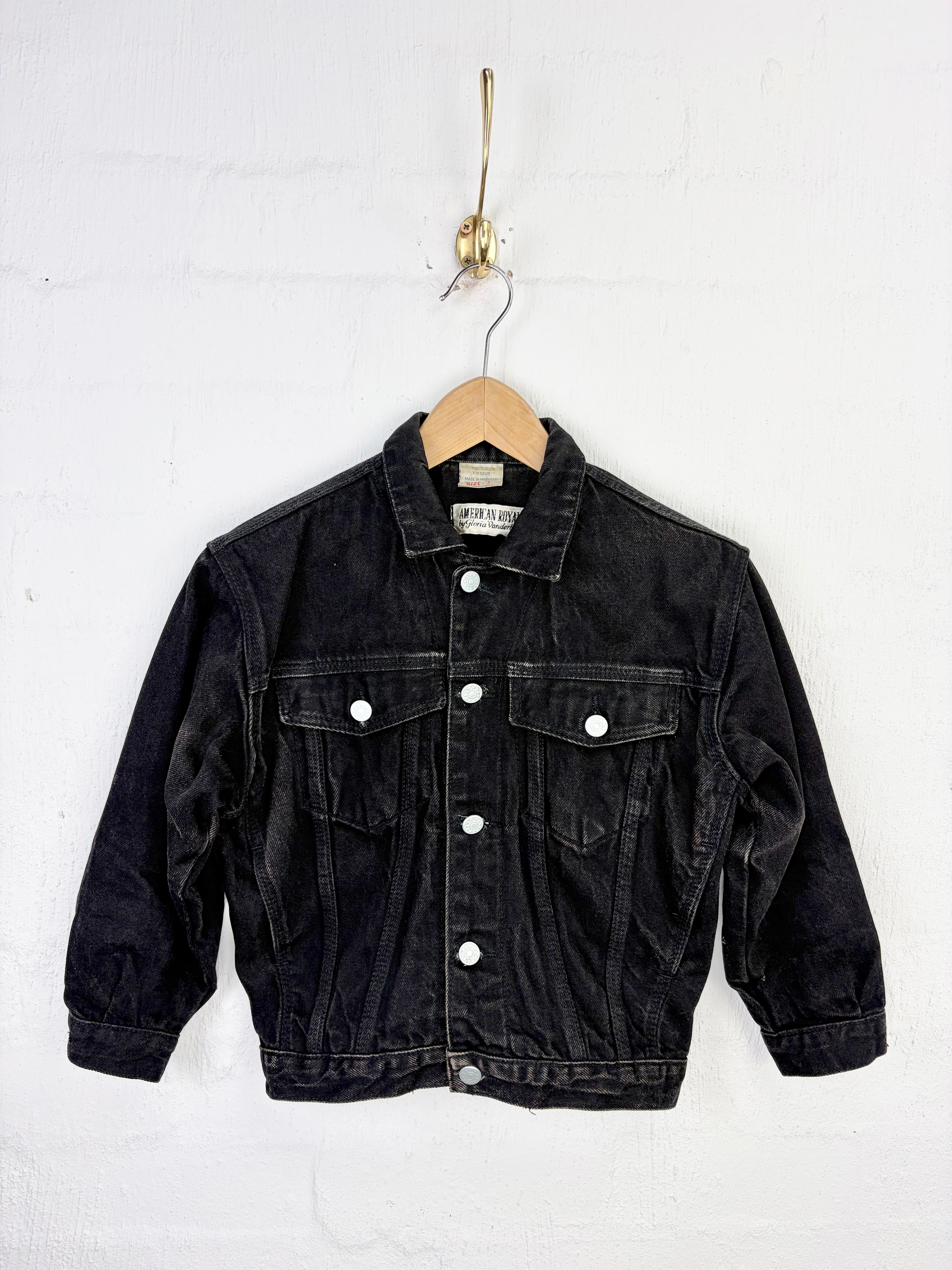A vintage black denim jacket hanging on a wooden hanger from a gold wall hook against a white painted brick wall.