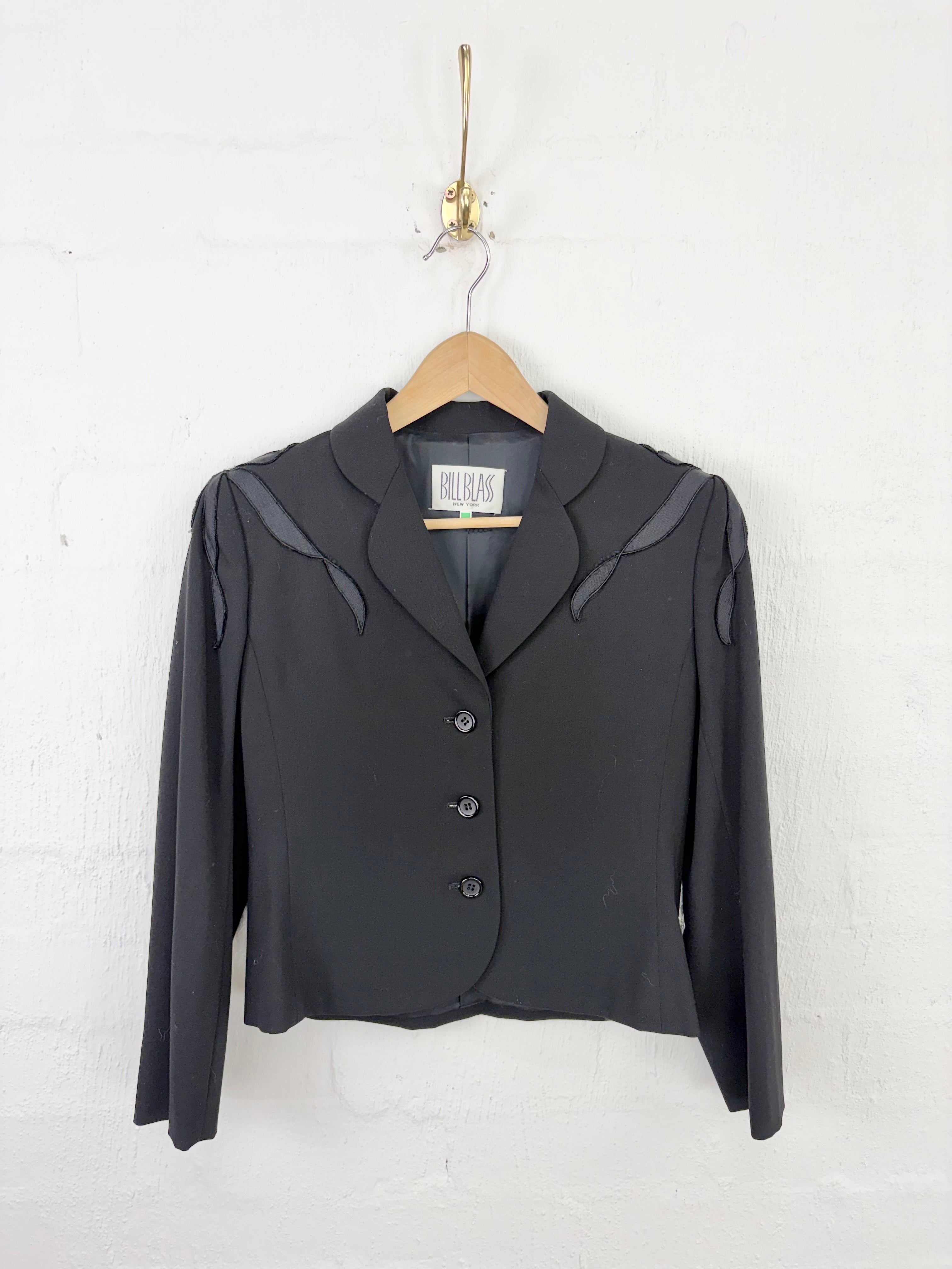 A black long-sleeve cropped blazer with notched lapels and dark shoulder detailing, hanging on a wooden hanger against a white painted brick wall.