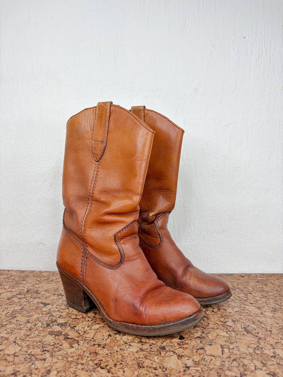 A pair of vintage brown leather cowboy boots with low block heels, displayed on a speckled stone surface against a white wall.