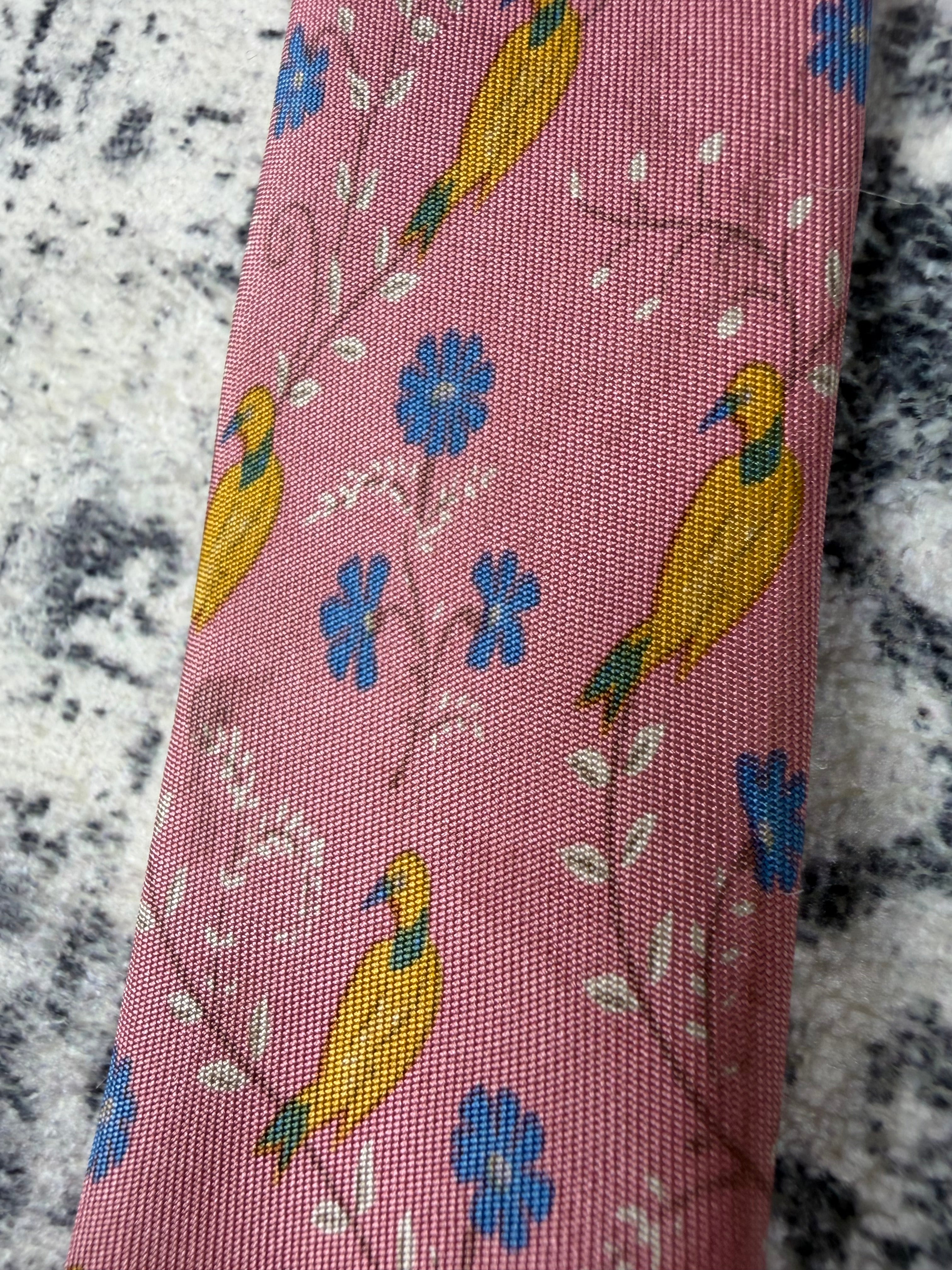 A close-up of a pink woven fabric with a repeating pattern of yellow birds and blue flowers, laid diagonally on a gray and white marbled surface.