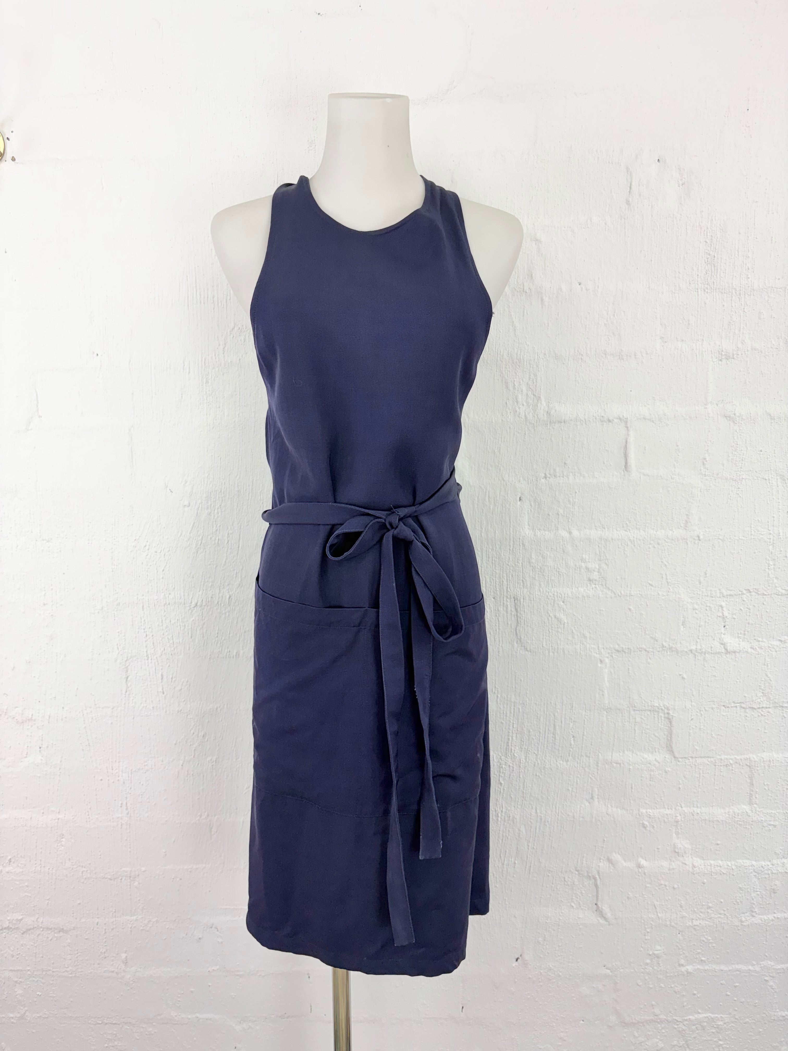 A sleeveless navy blue apron dress displayed on a white mannequin torso against a white painted brick wall. The dress features a tie waist belt and front pockets.