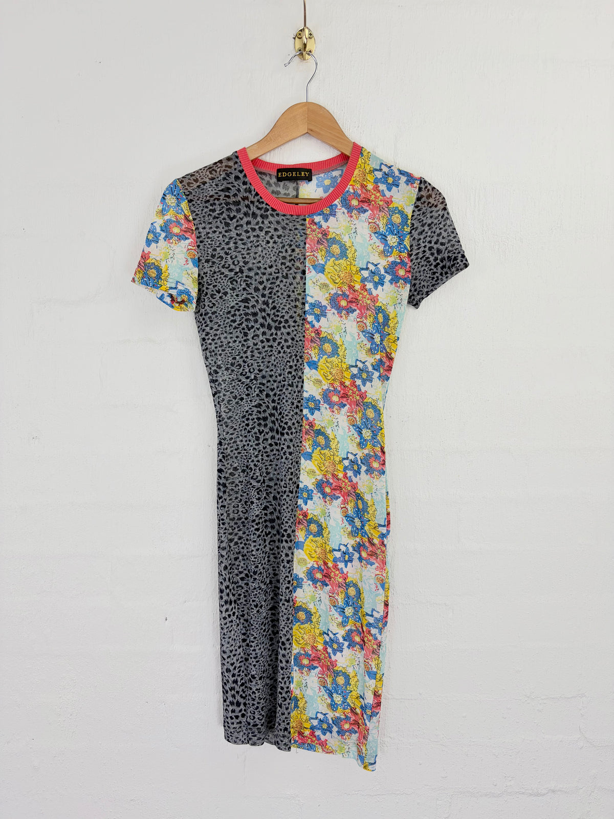 A short-sleeve dress hanging on a wooden hanger against a white brick wall. The dress features a split design: one side has a black leopard print, and the other has a colorful floral print with blue, yellow, red, and white flowers.