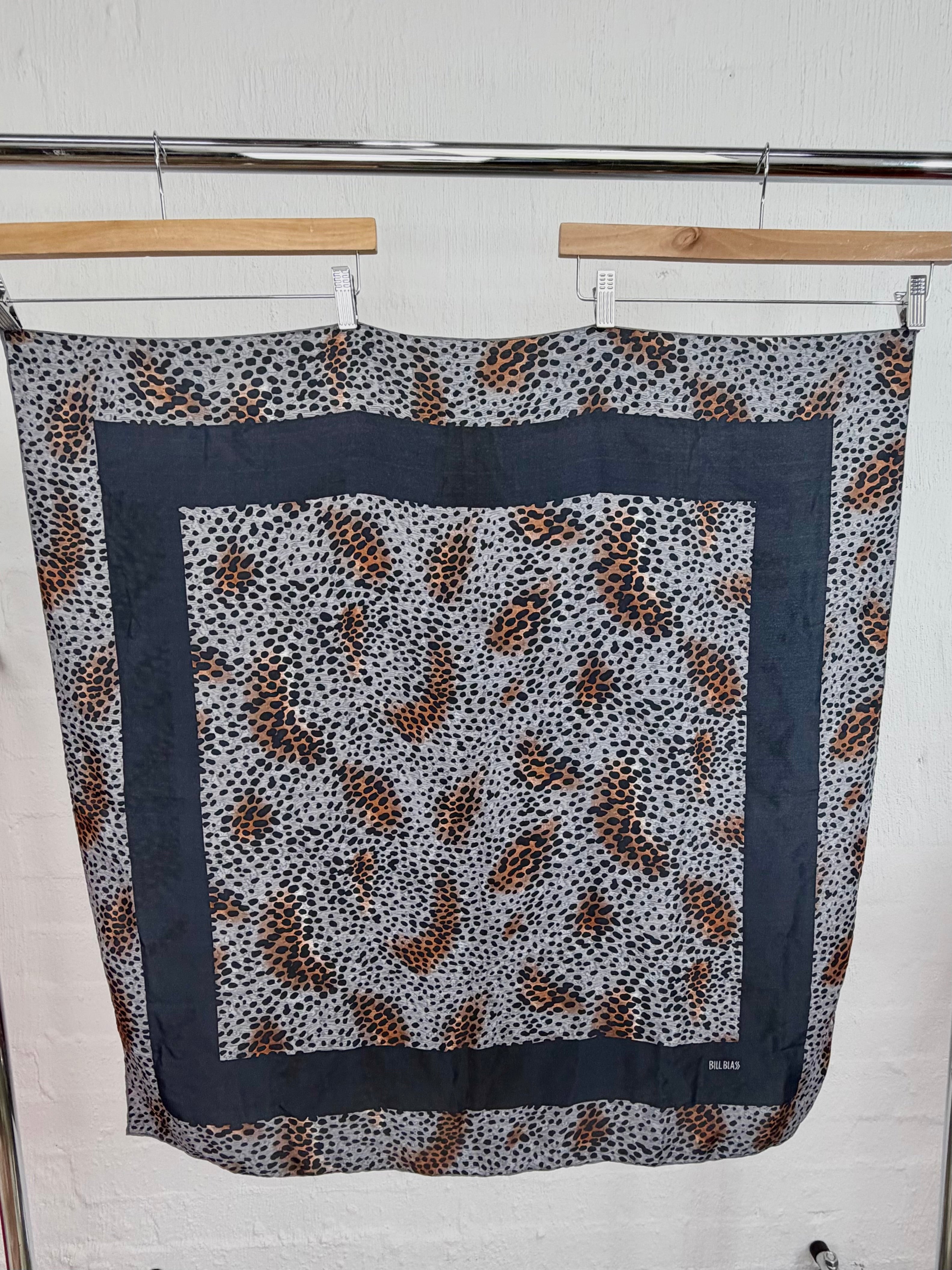 A square scarf with a leopard print pattern in brown, black, and white, featuring a wide dark gray border, hanging on wooden hangers against a white brick wall.