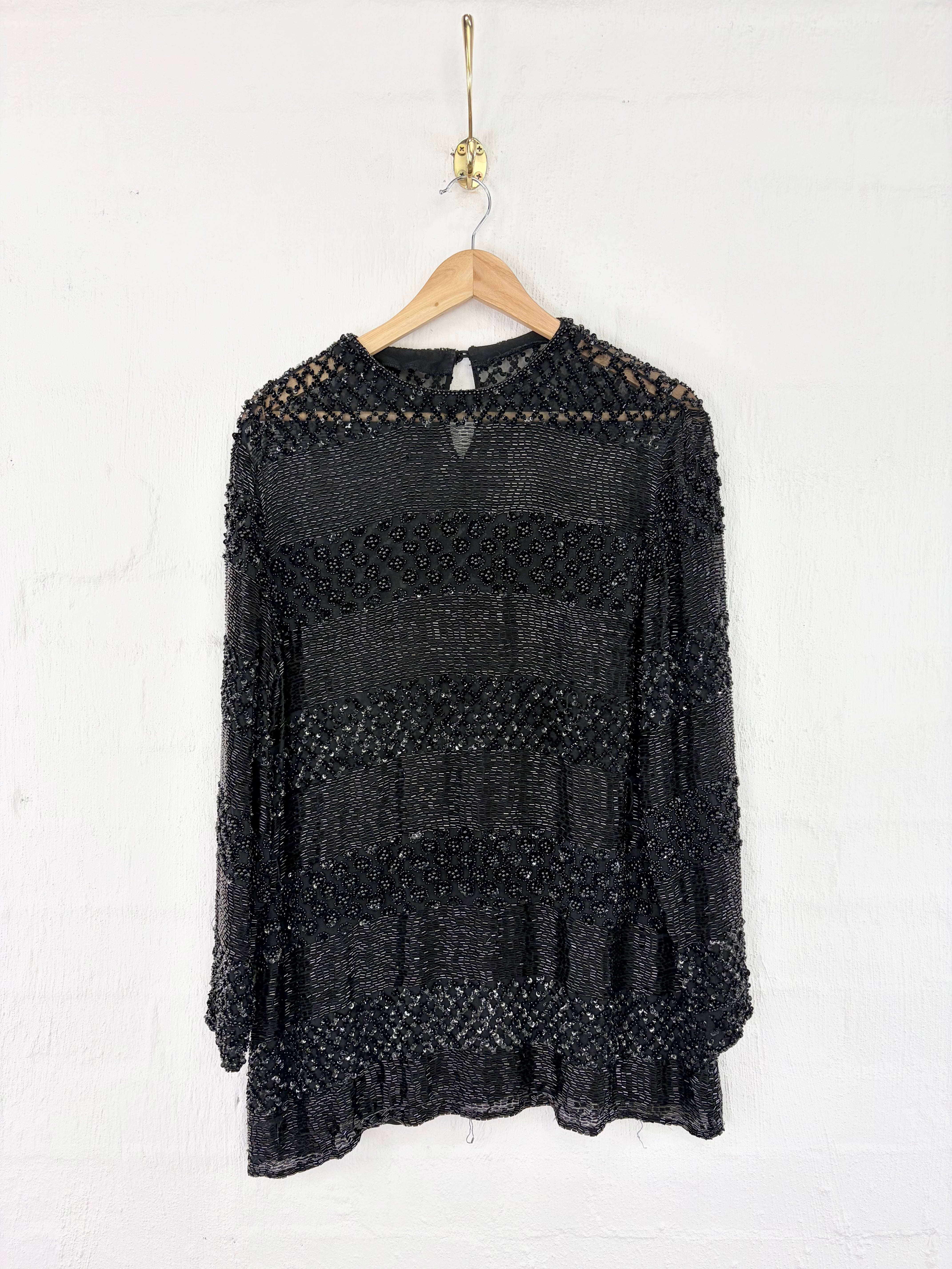 A black textured knit long-sleeve top with a crew neck, hanging on a wooden hanger against a white painted brick wall.