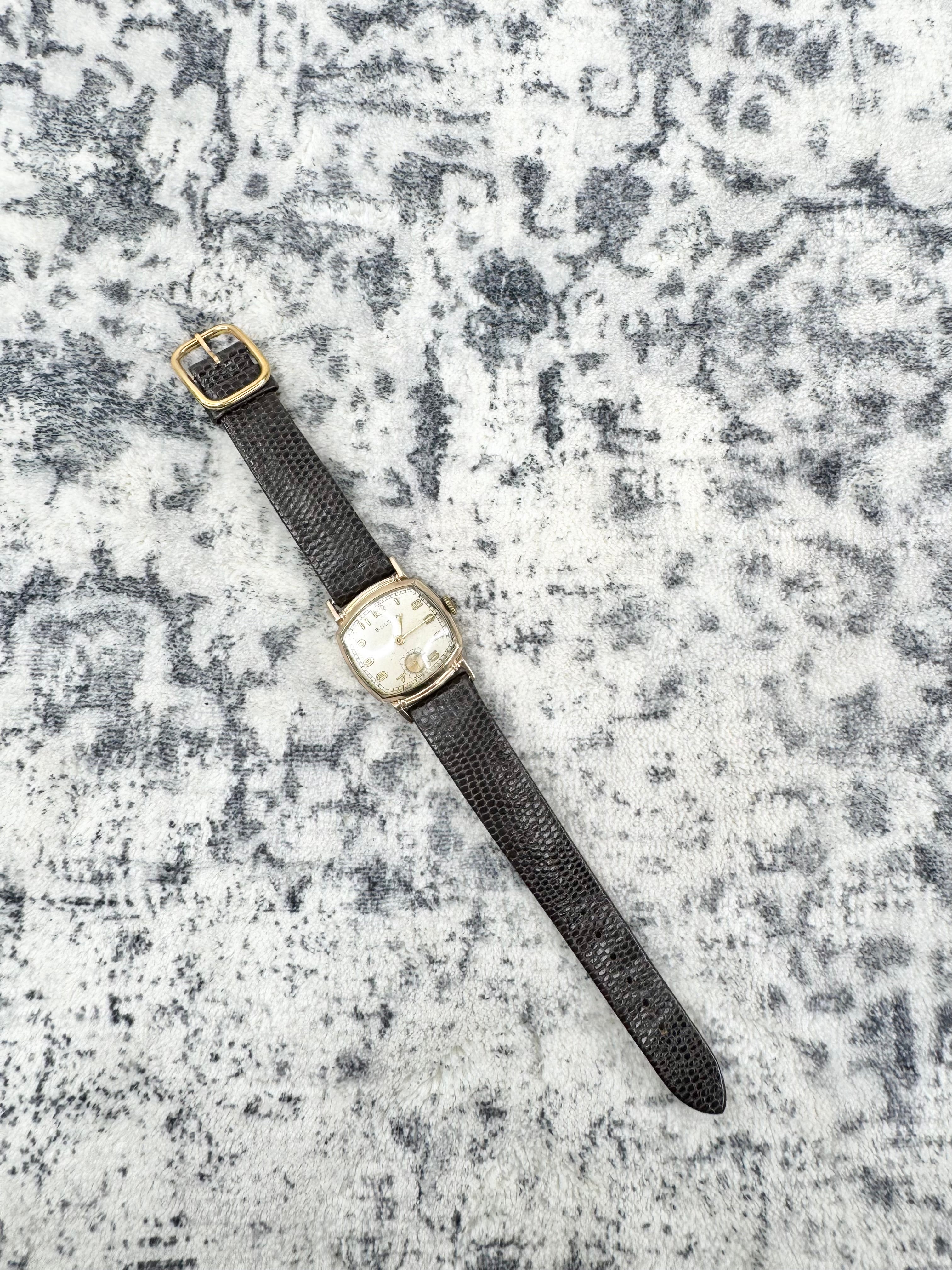 A vintage-style wristwatch with a textured dark gray strap, gold-tone case, and cream-colored dial, laid diagonally on a mottled gray and white textured surface.