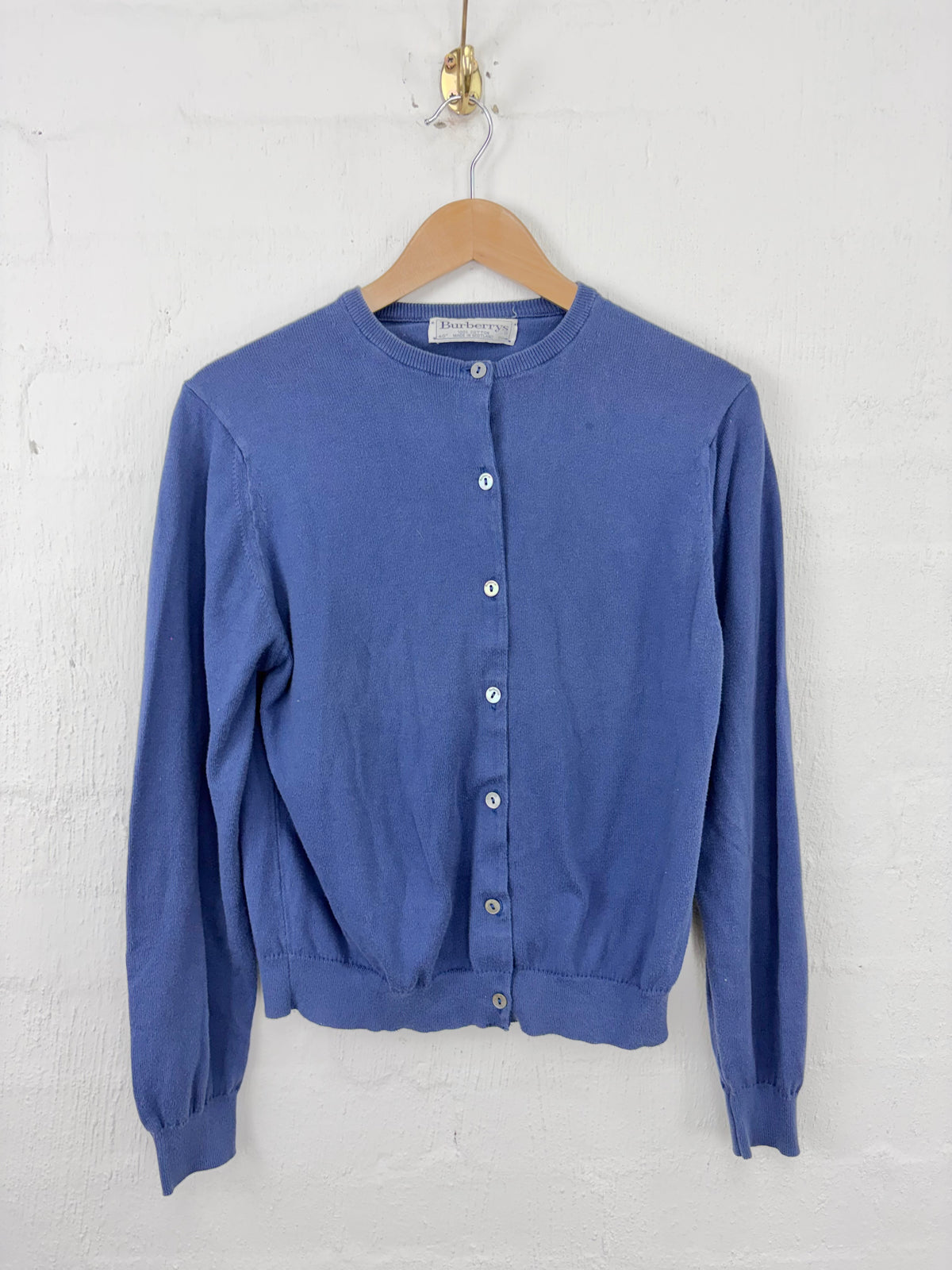 A blue long-sleeve button-front cardigan sweater hanging on a wooden hanger against a white painted brick wall.