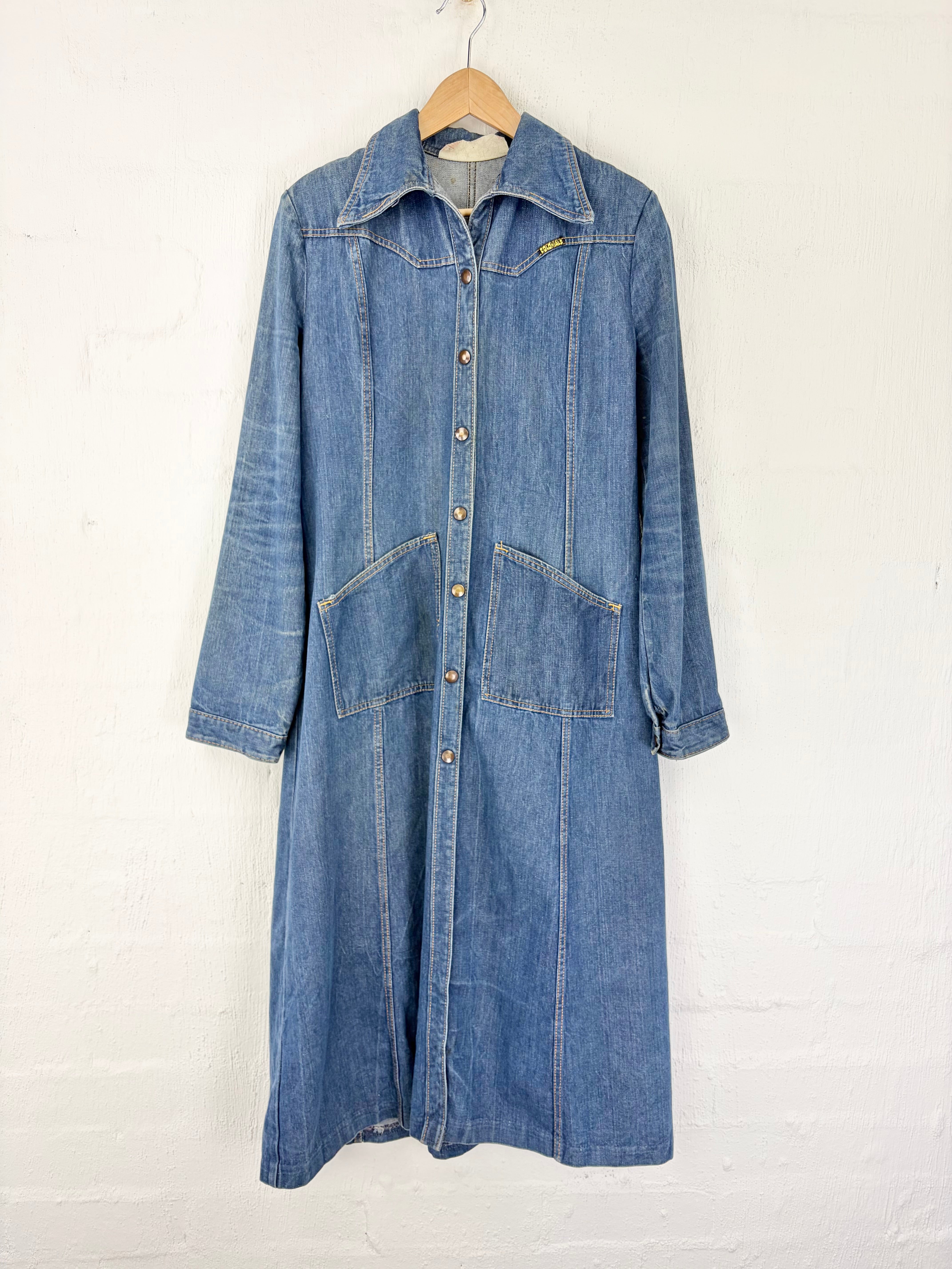 A blue denim longline shirt jacket with a button-down front, collar, and front patch pockets, hanging on a wooden hanger against a white painted brick wall.