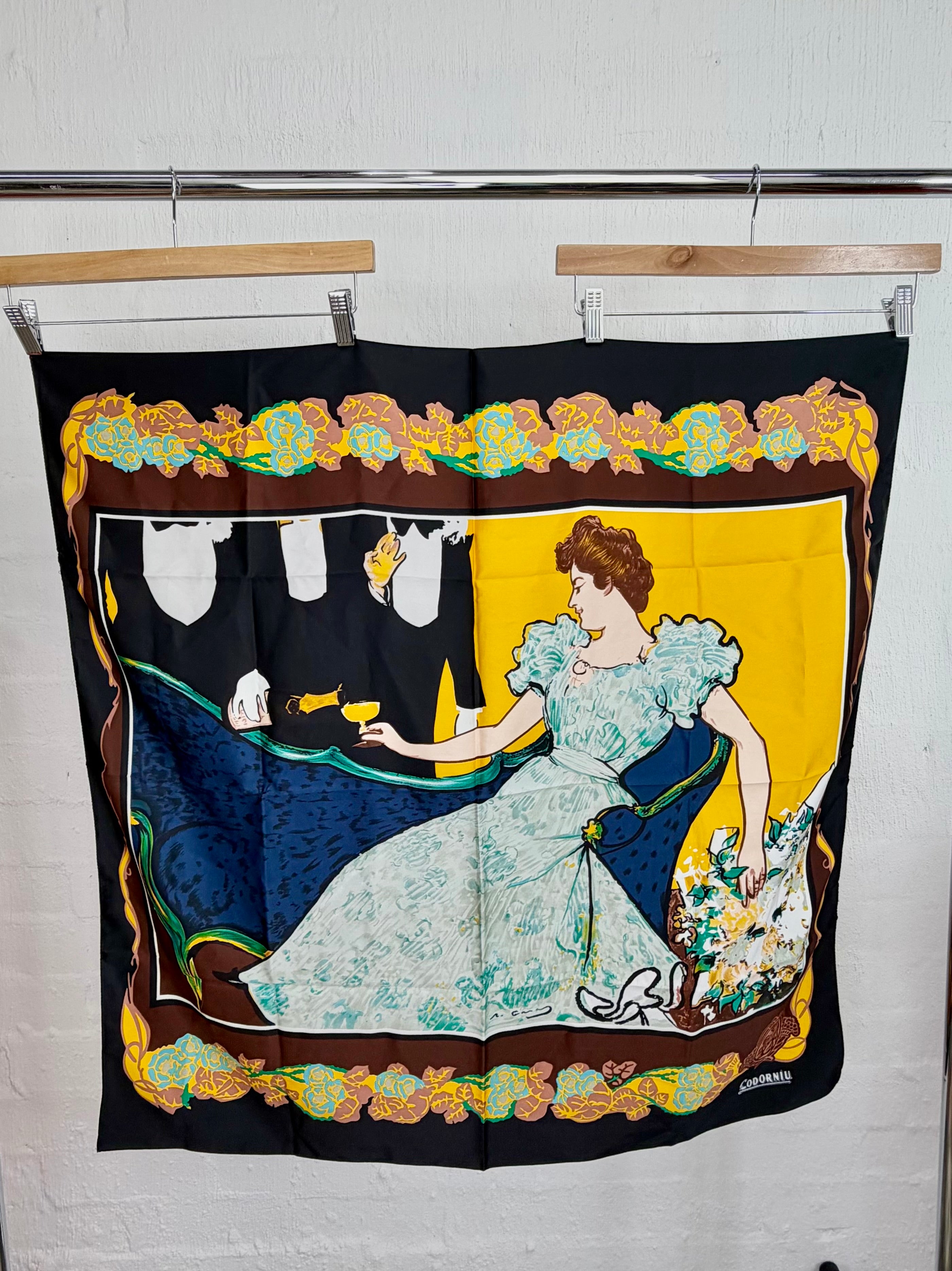 A square silk scarf featuring a vintage Art Nouveau illustration of a woman in a light blue gown on a yellow and black background, hanging from wooden hangers on a metal rod against a white brick wall.