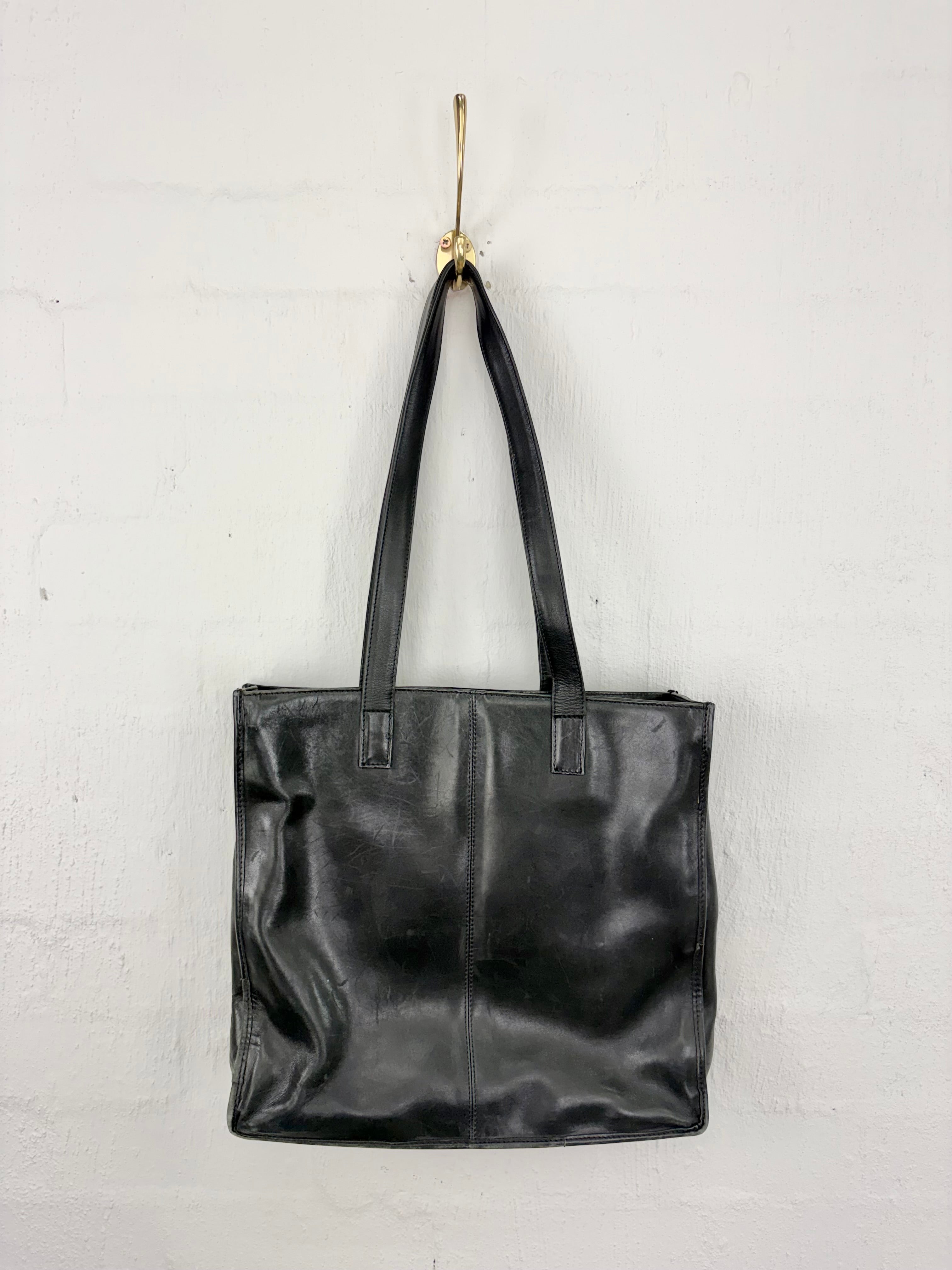 A black leather tote bag hanging from a gold hook on a white textured wall.