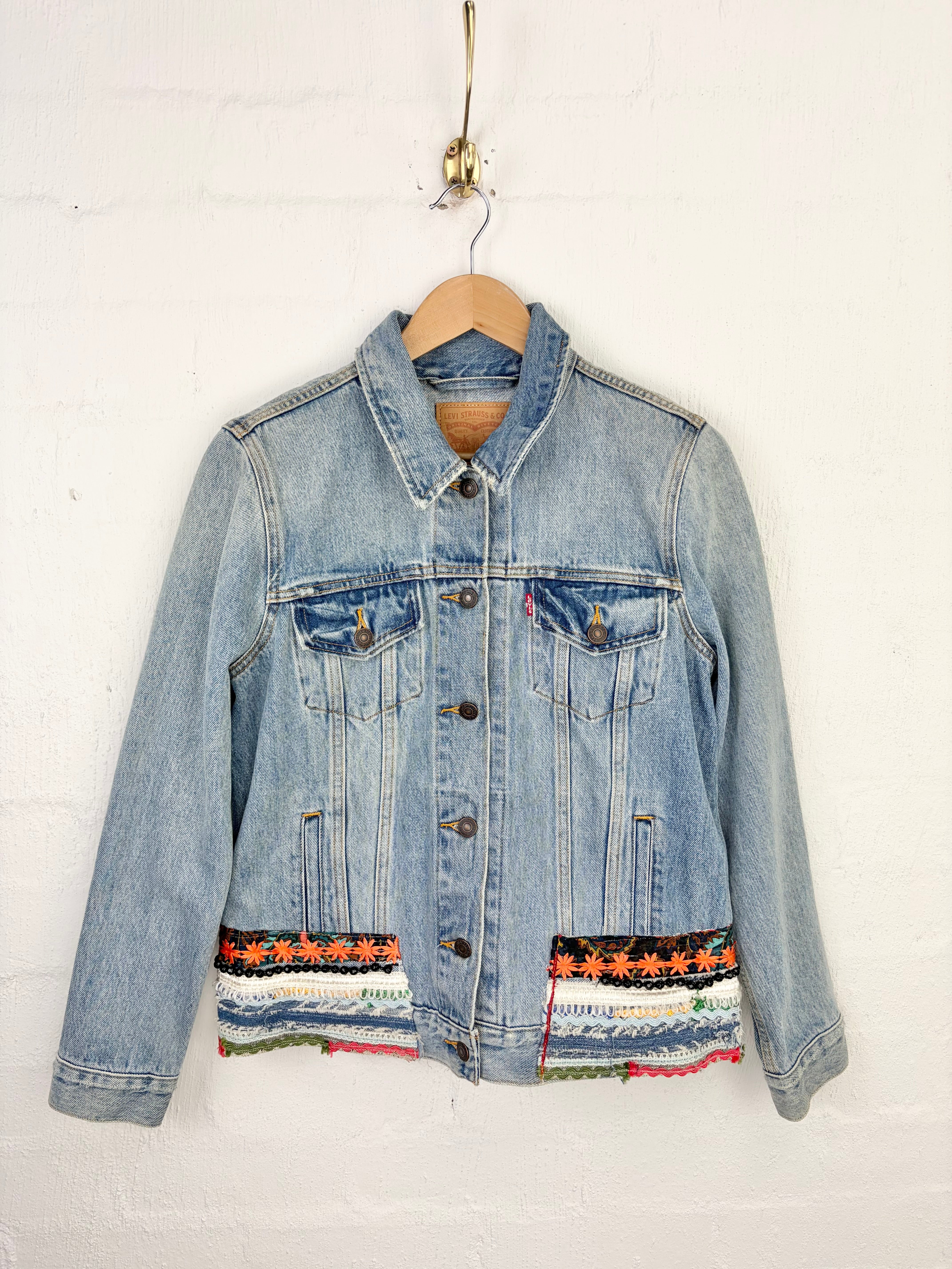 A light blue denim jacket with colorful patterned fabric trim along the hem and pocket flaps, hanging on a wooden hanger against a white painted brick wall.