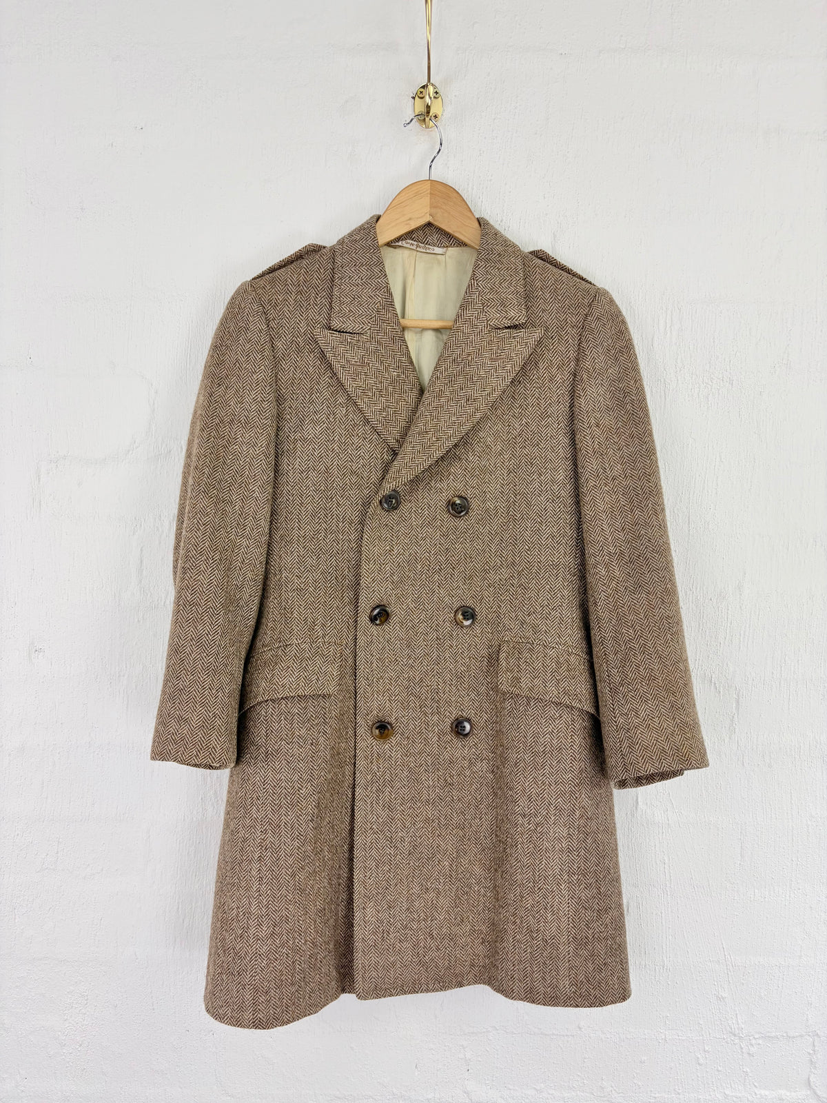 A classic double-breasted wool overcoat in a textured taupe-brown tweed, hanging on a wooden hanger against a white painted brick wall.