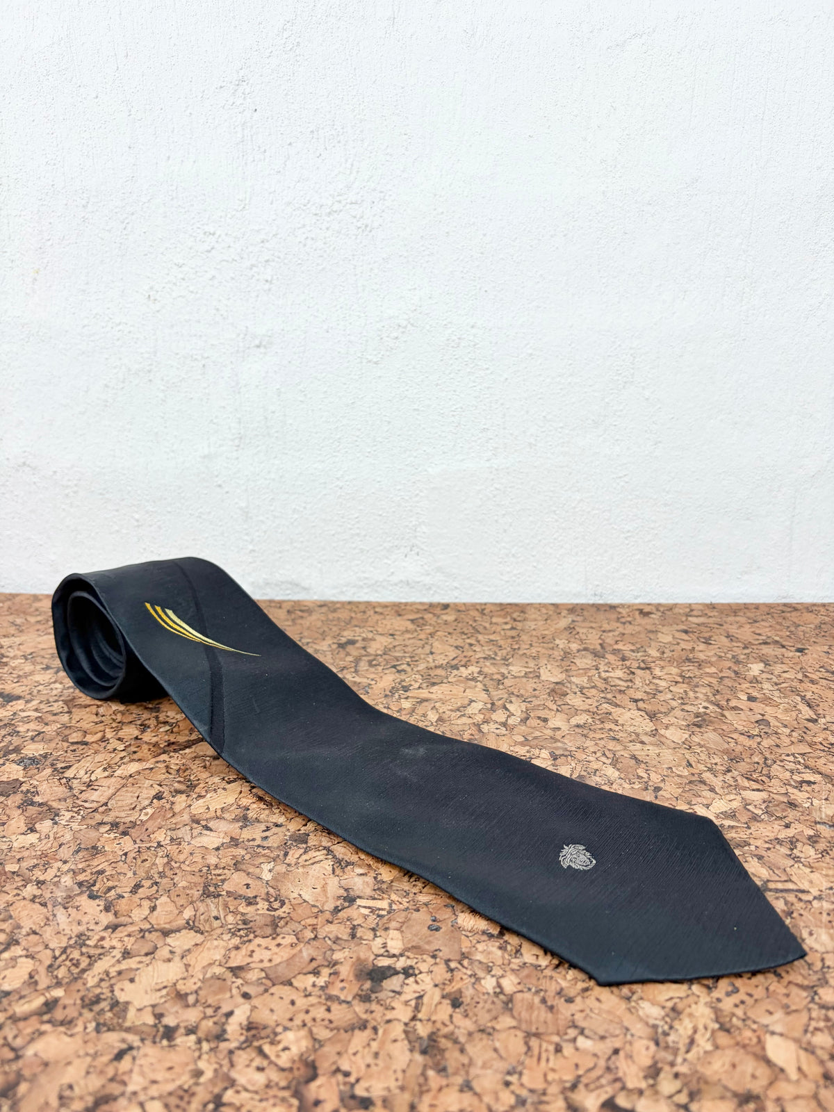 A black necktie with a gold wave pattern on the left side and a small silver crest on the tip, displayed on a cork surface against a white wall.
