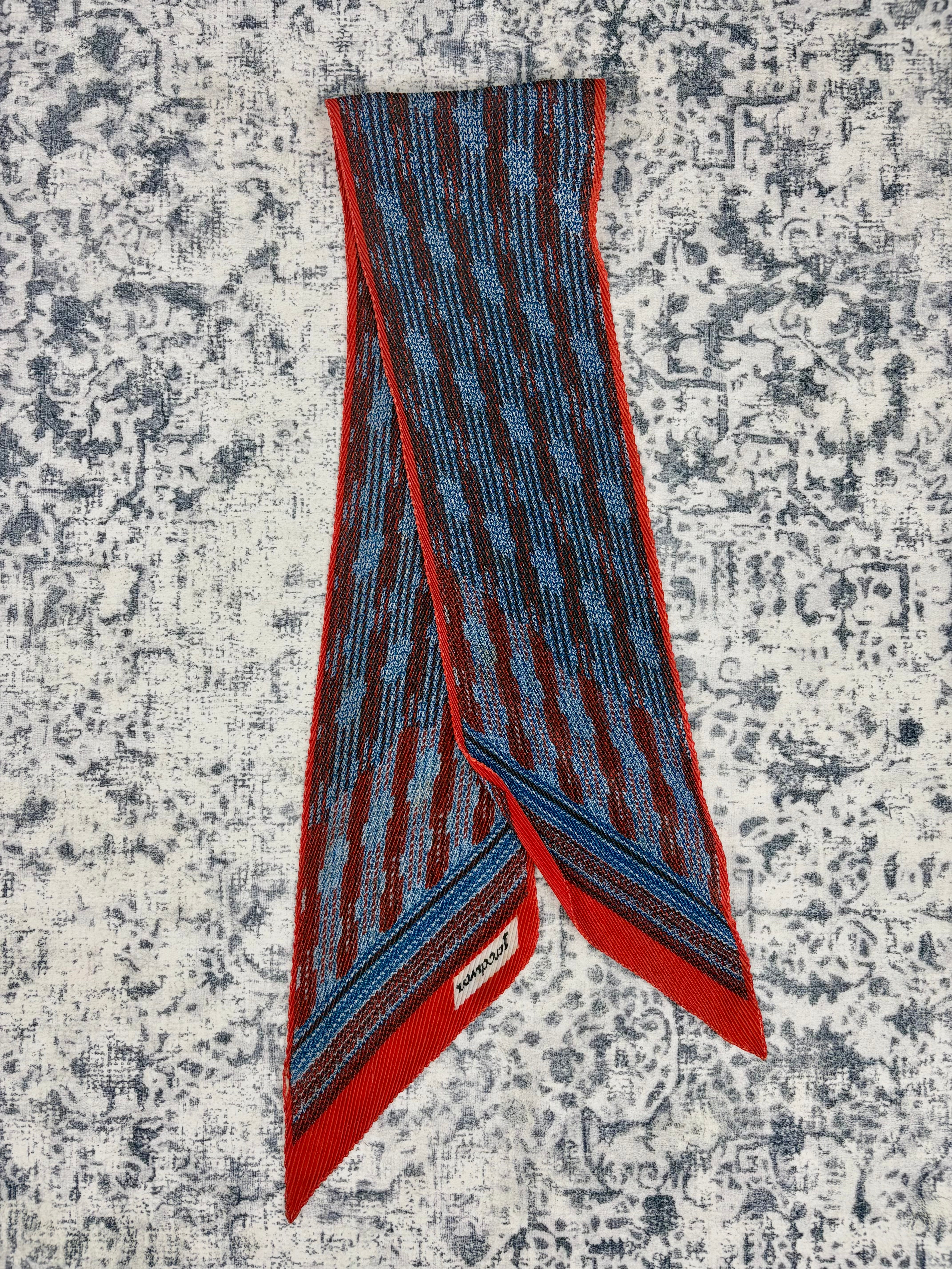 A folded patterned scarf with red, blue, and black stripes, displayed on a distressed gray and white patterned background. The brand name 'SARABAND' is visible on a small white label near the bottom corner.