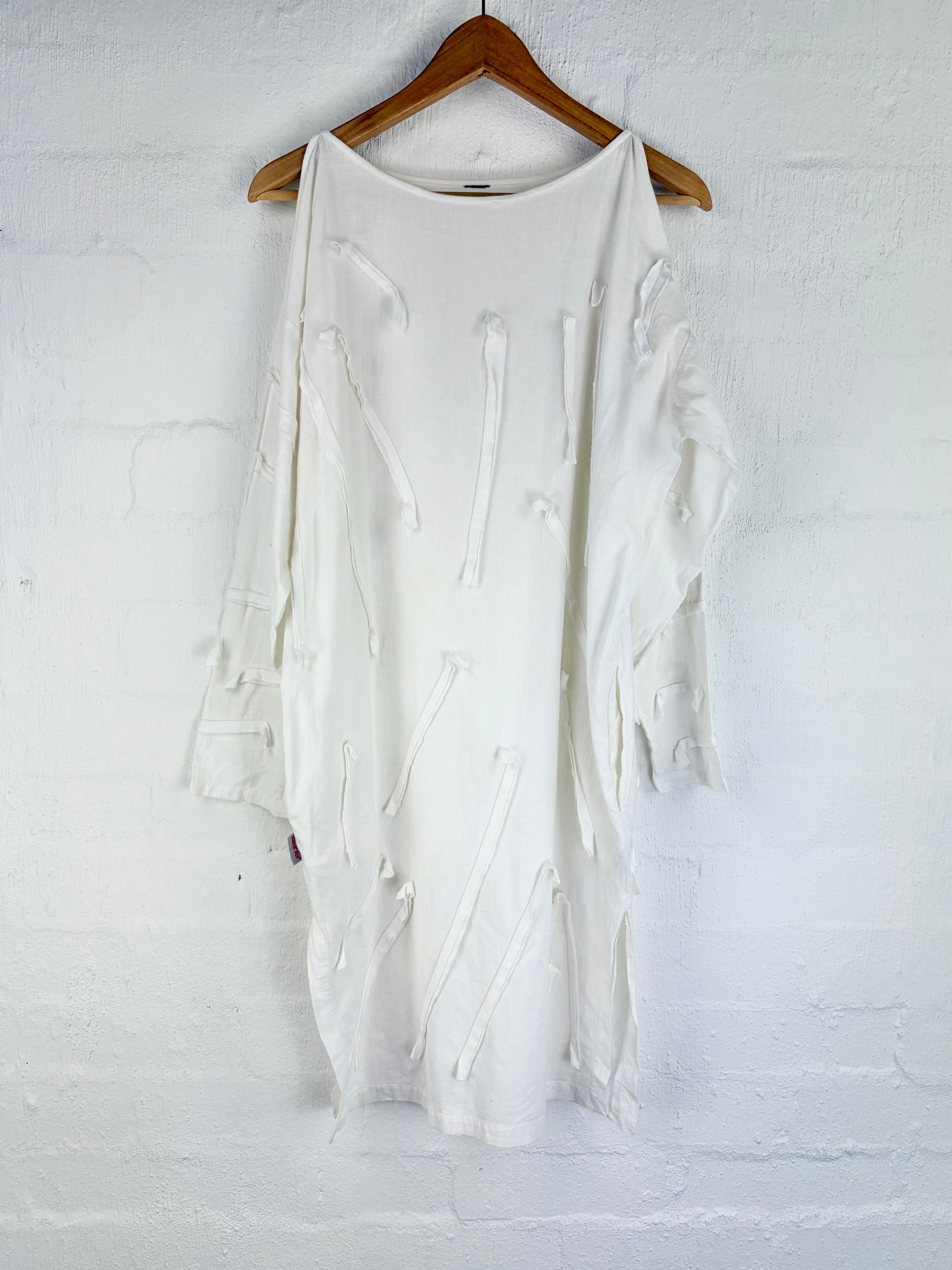 An off-white, distressed long-sleeve tunic top hanging on a wooden hanger against a white painted brick wall.