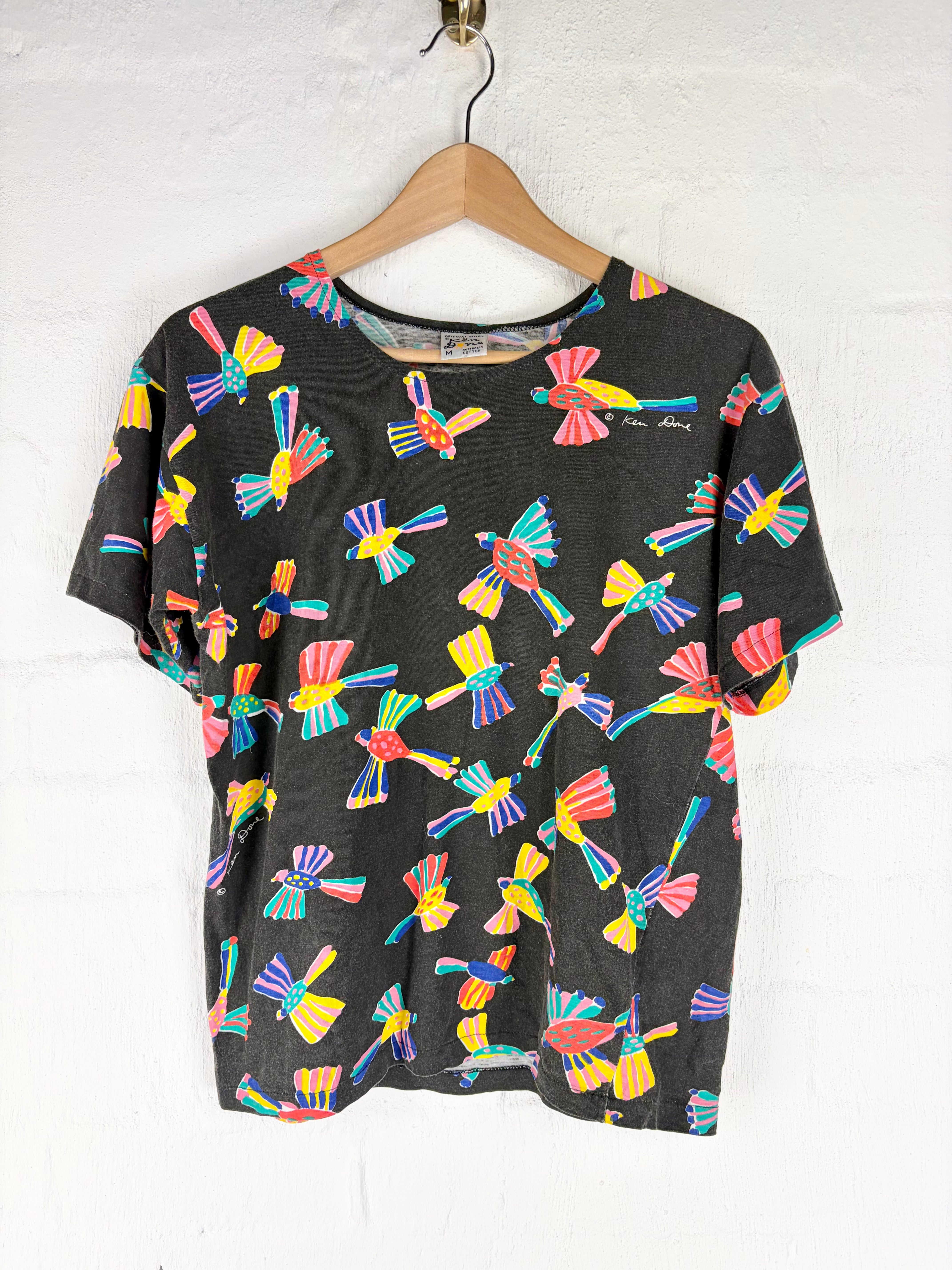 A black short-sleeve t-shirt with a colorful dragonfly pattern, hanging on a wooden hanger against a white textured wall.