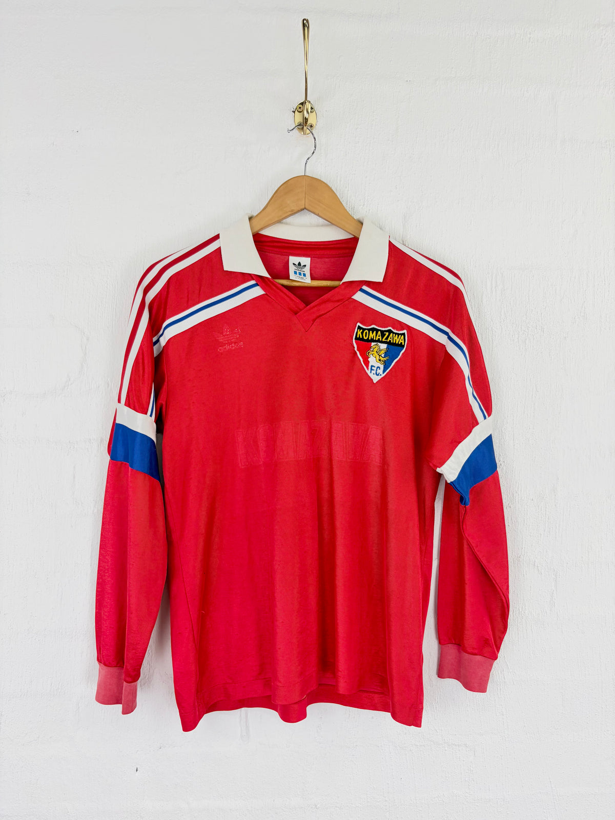 A vintage red long-sleeve soccer jersey with white and blue stripes on the shoulders and sleeves, hanging on a wooden hanger against a white brick wall. The jersey features a white collar, a yellow and blue club crest on the left chest, and an Adidas logo on the right chest.