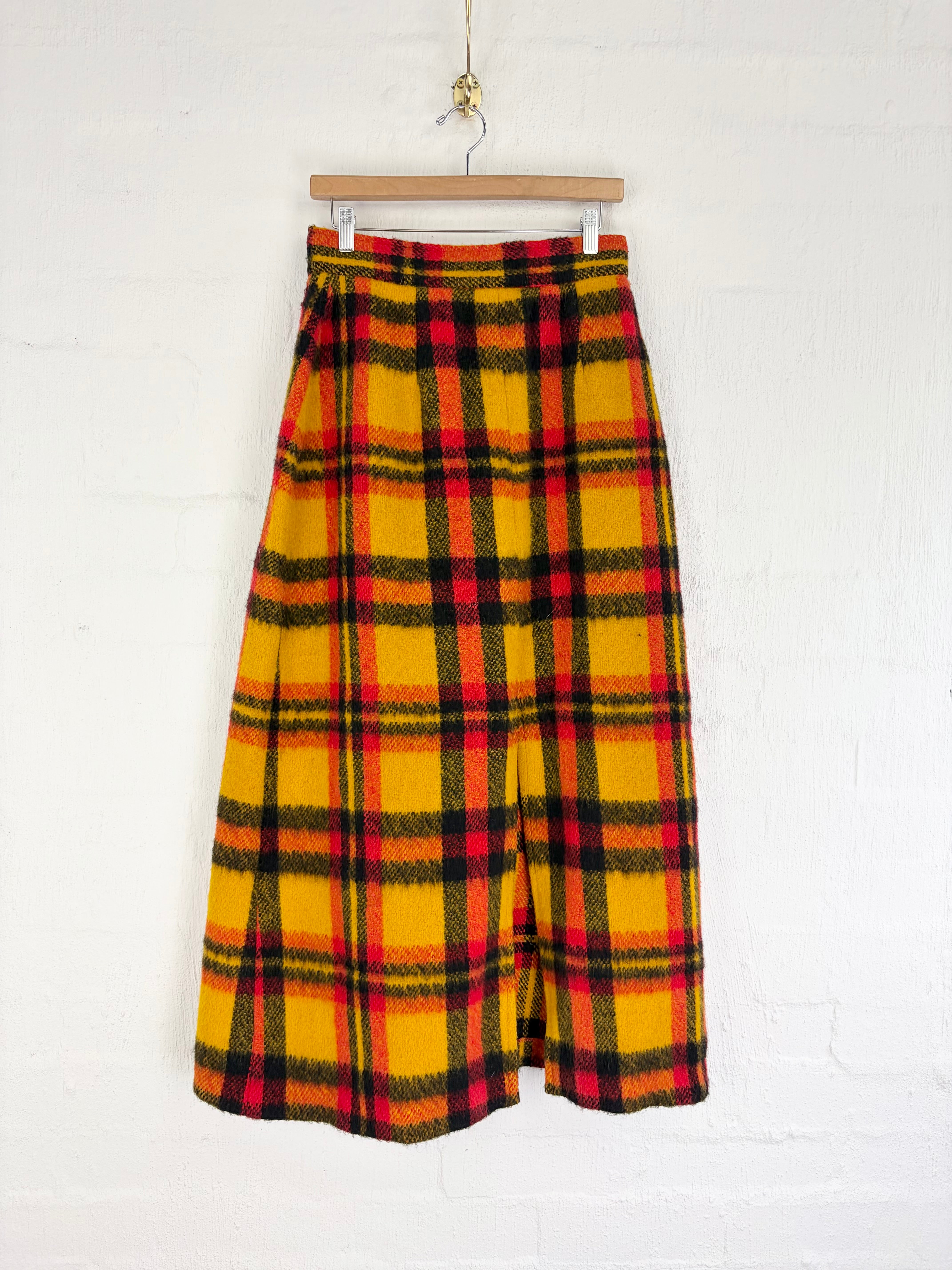 A yellow wool plaid skirt with red and black stripes, hanging on a wooden hanger against a white brick wall.