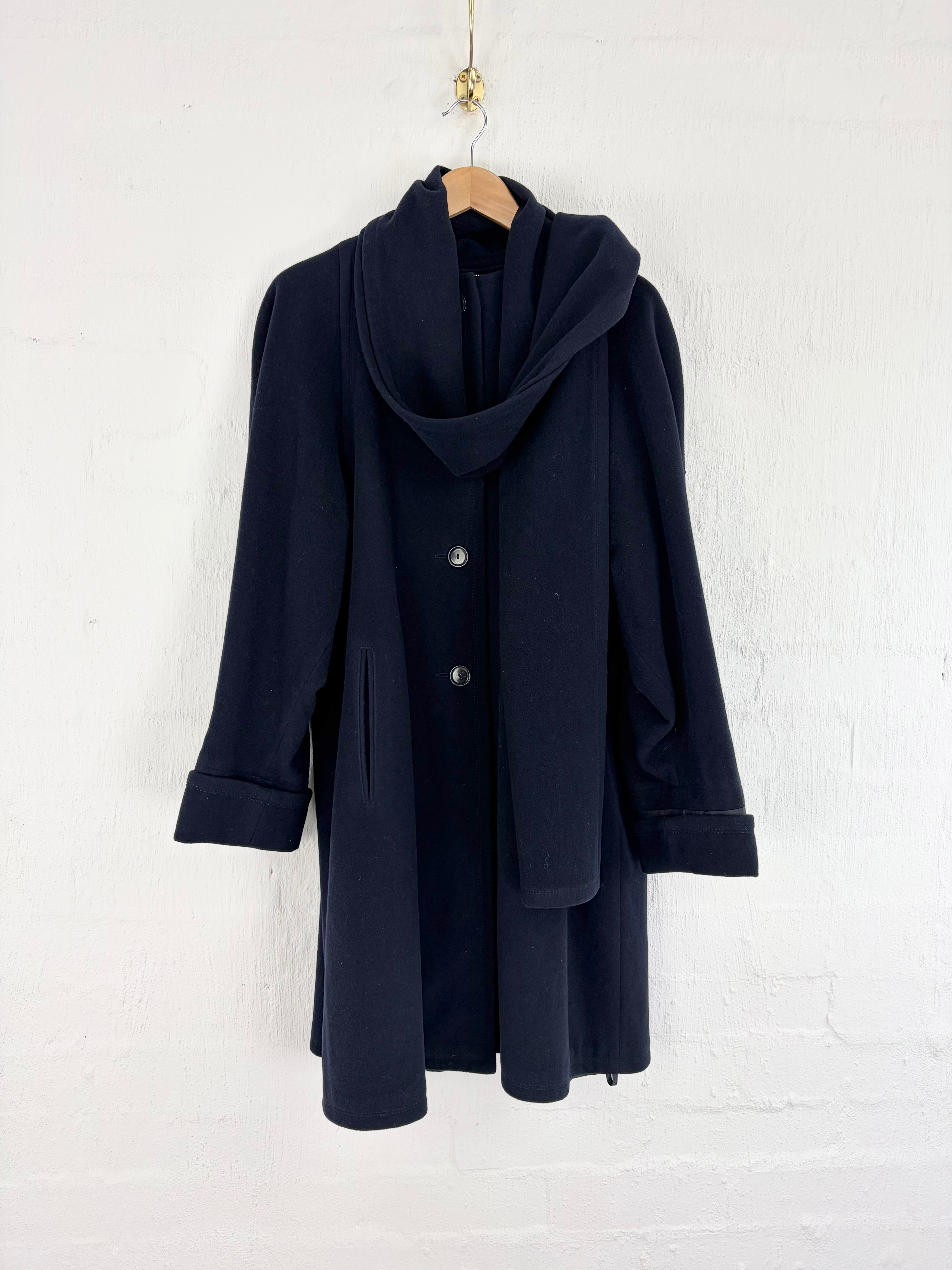 A dark navy blue coat with a draped, oversized cowl collar and front buttons, hanging on a wooden hanger against a white painted brick wall.
