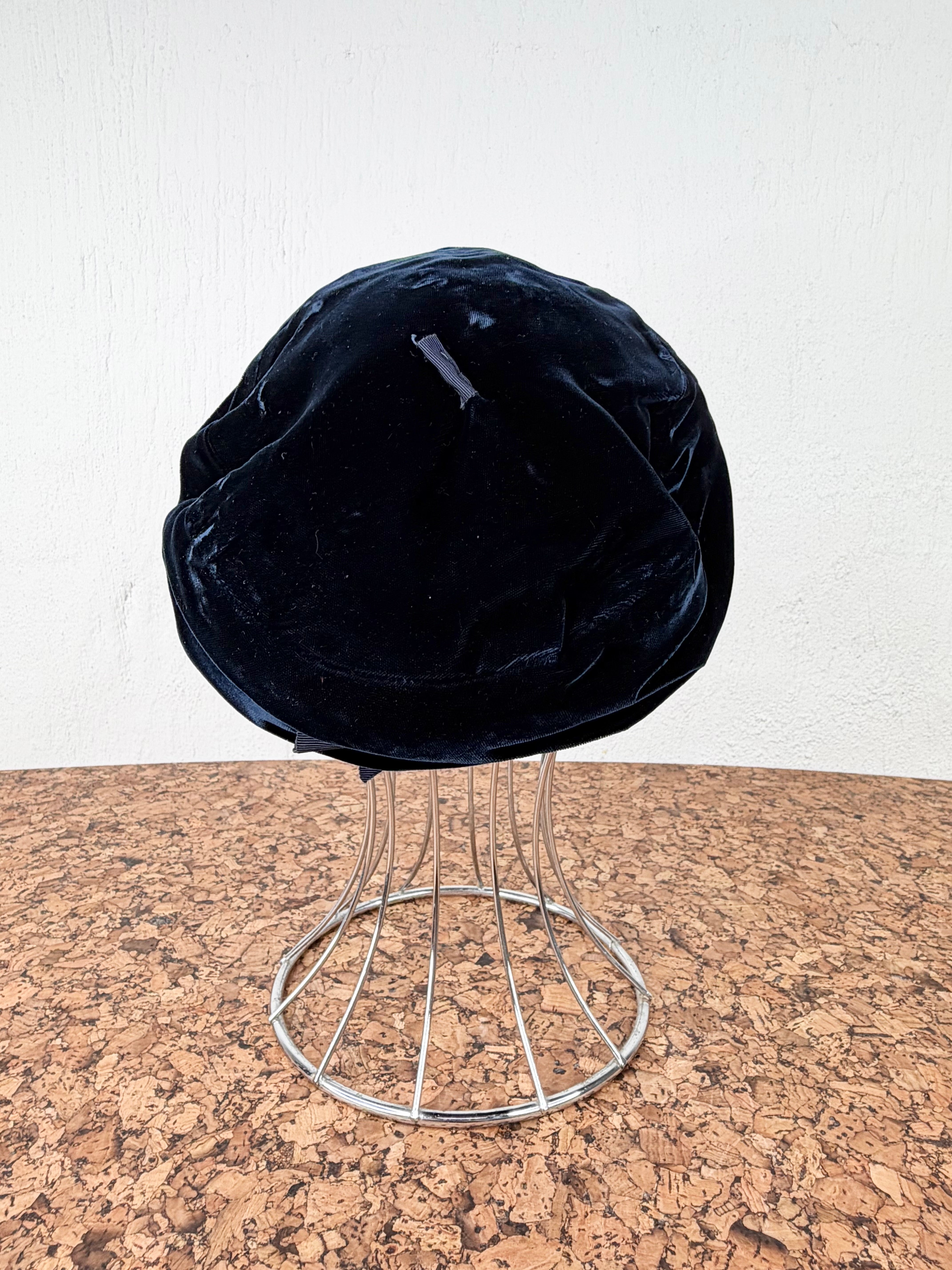A black velvet beret-style hat displayed on a silver metal wire stand against a white wall and cork tabletop.