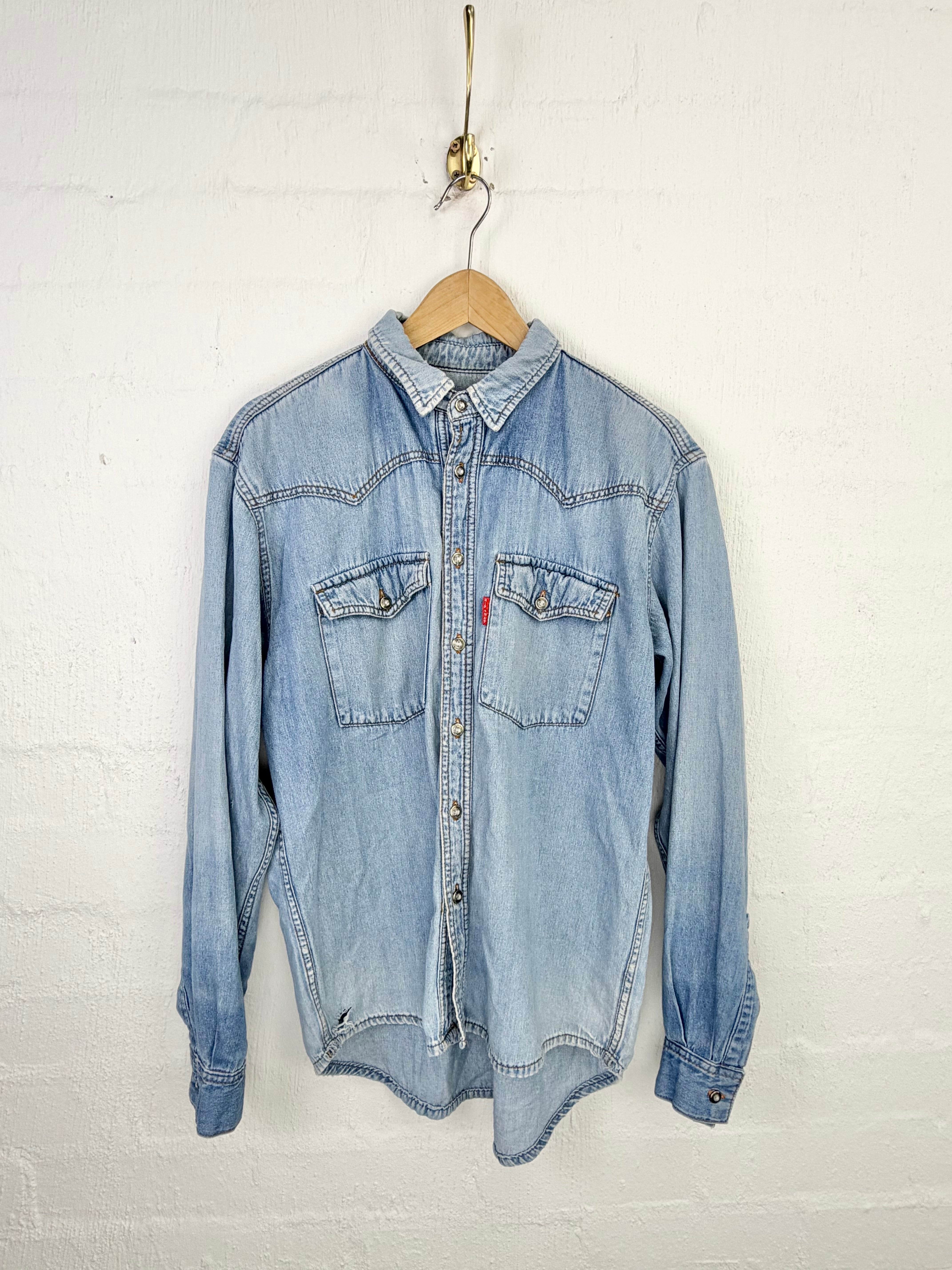 A light blue denim western-style shirt jacket hanging on a wooden hanger against a white painted brick wall.