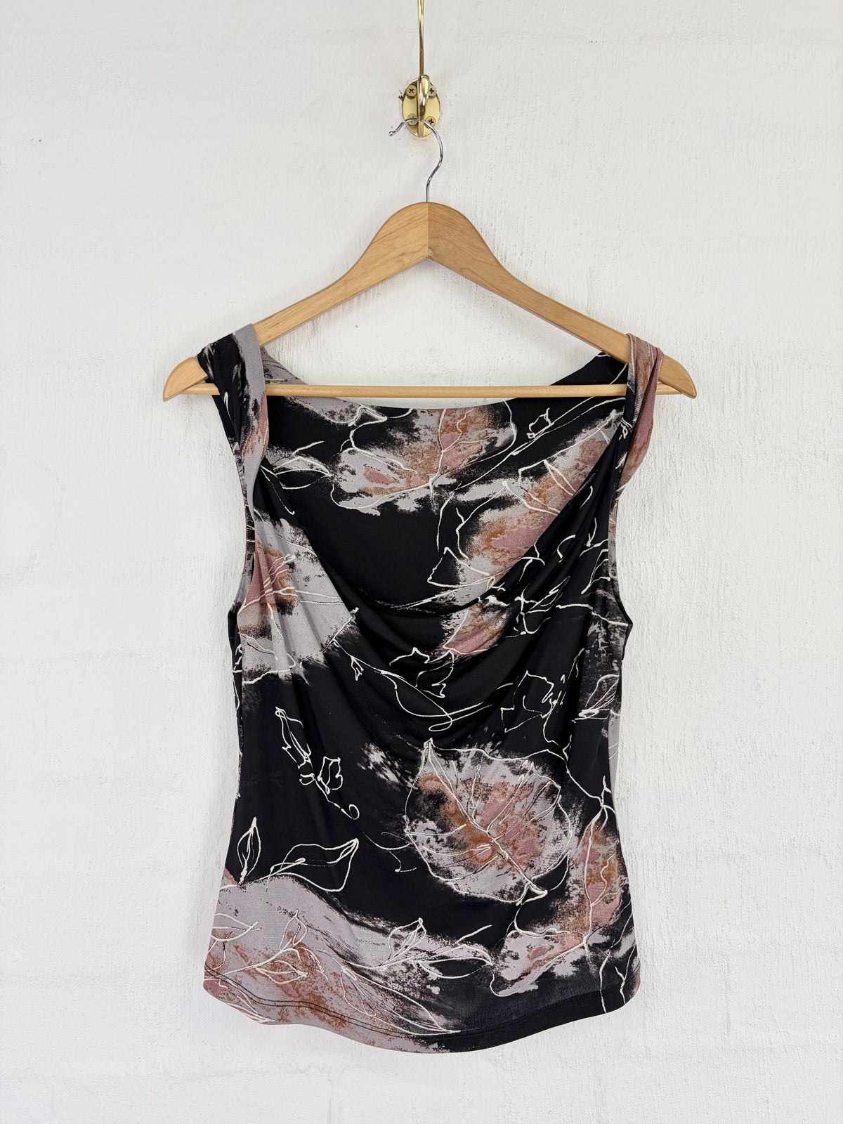 A sleeveless draped tank top with a black base and abstract floral print in dusty pink, beige, and white, hanging on a wooden hanger against a white brick wall.