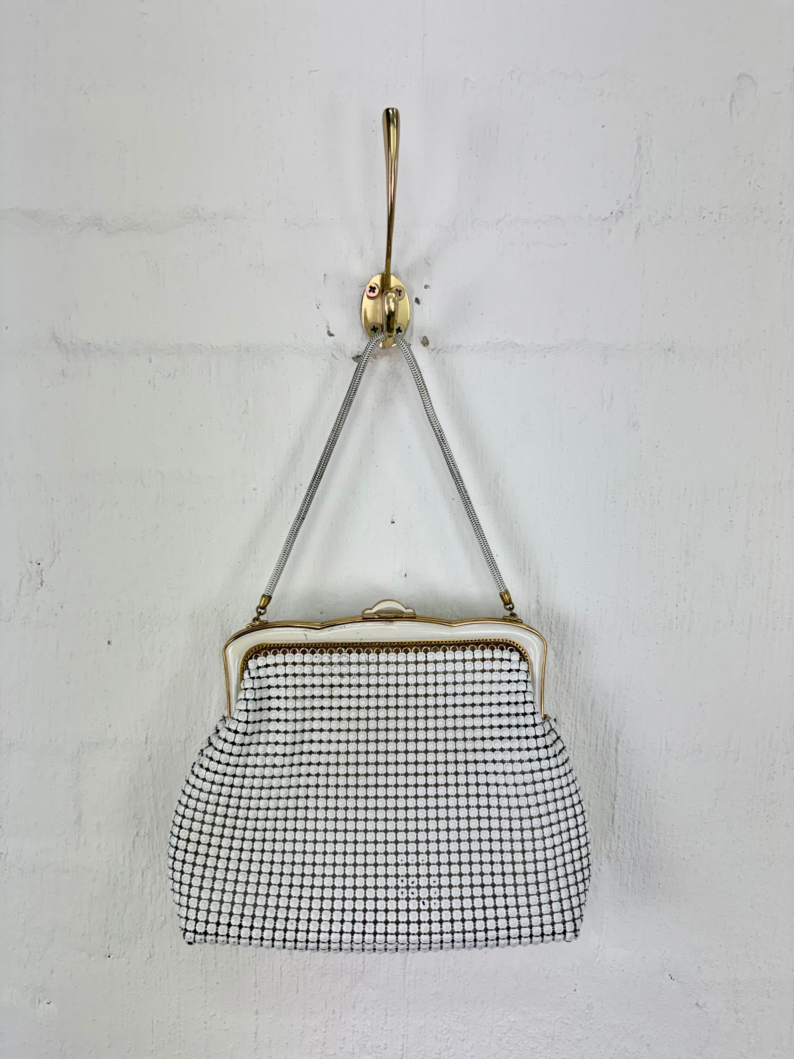 A vintage-style black and white checkered clutch purse with a gold-tone frame and strap, hanging from a gold hook against a white textured wall.