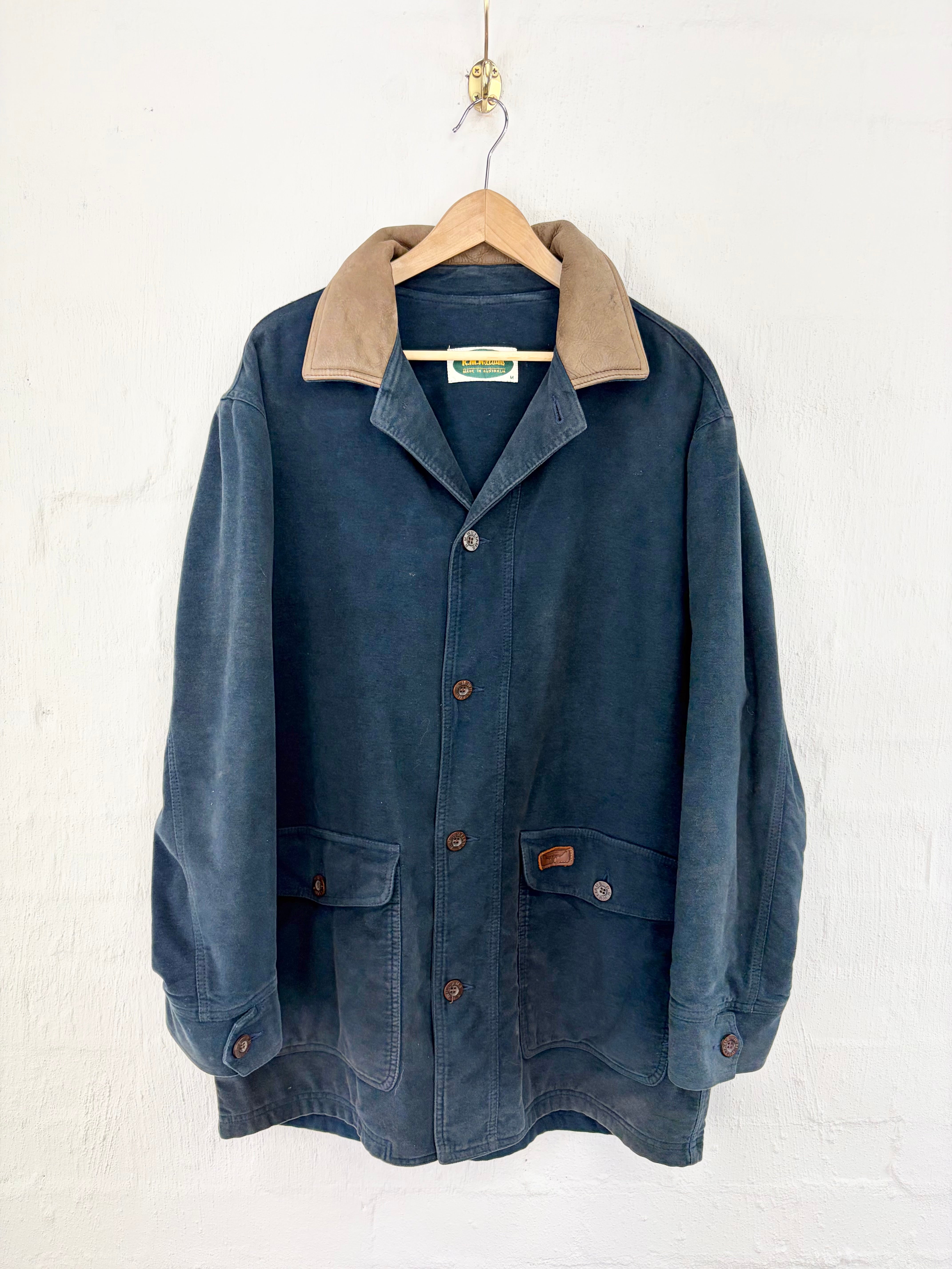 A vintage dark blue corduroy jacket with a tan corduroy collar, hanging on a wooden hanger against a white painted brick wall.