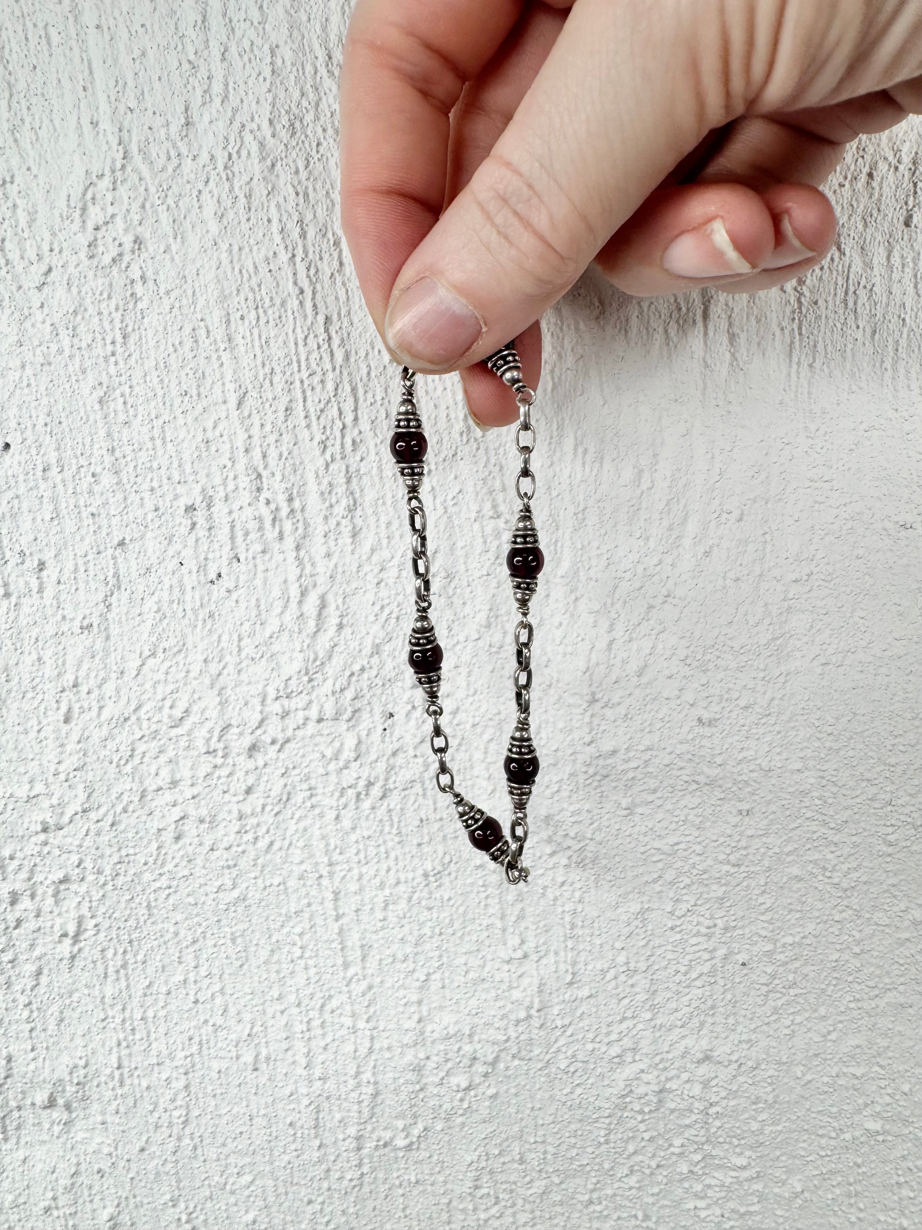 A close-up of a hand holding a delicate silver-toned chain necklace with dark, bead-like accents against a textured white wall.