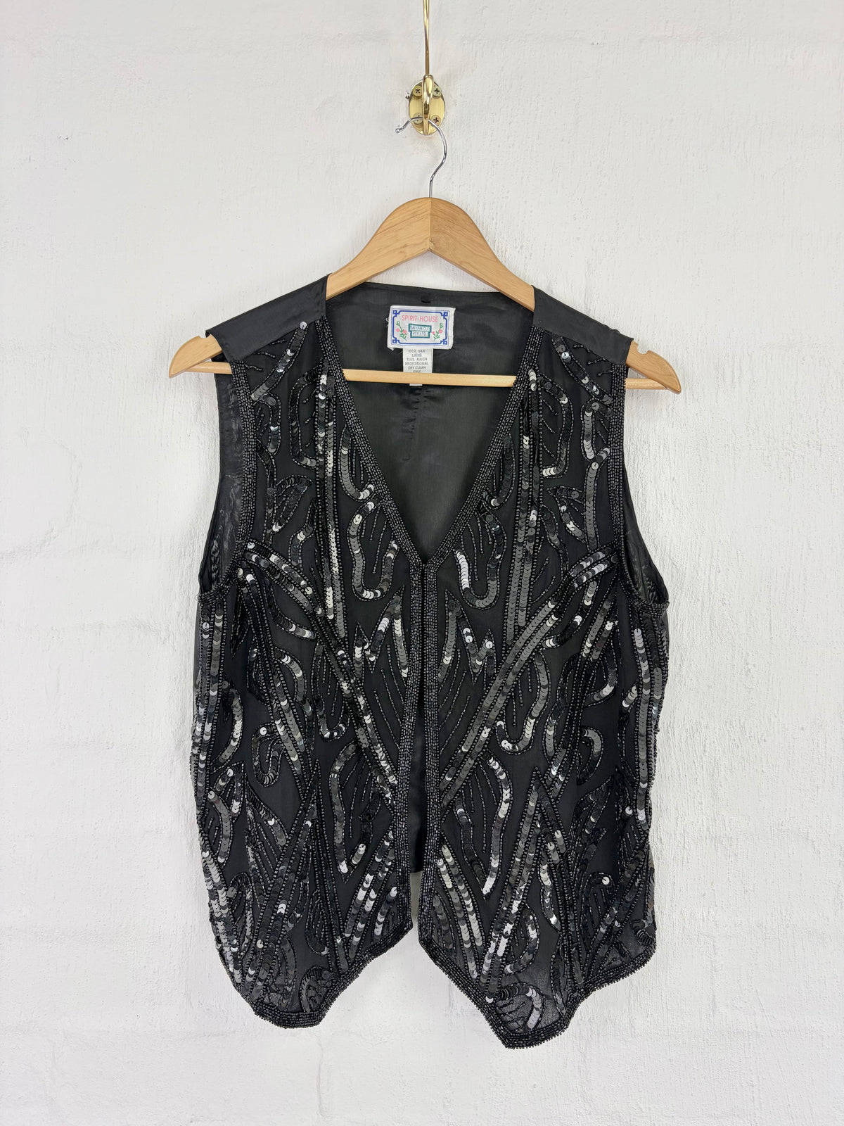 A black sleeveless vest with silver sequin beadwork, hanging on a wooden hanger against a white painted brick wall.
