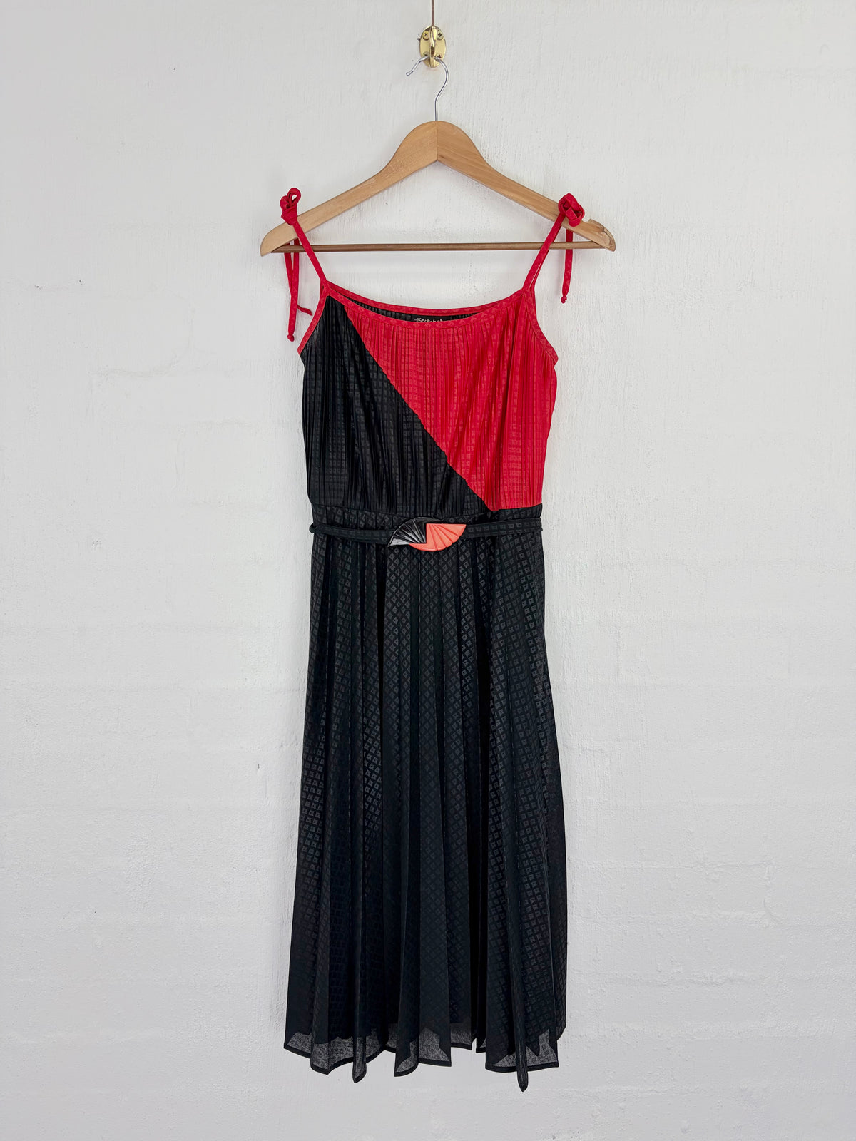 A sleeveless midi dress with thin shoulder ties, featuring a diagonal color-block design with a textured red upper panel and a black dotted skirt, hanging on a wooden hanger against a white brick wall.