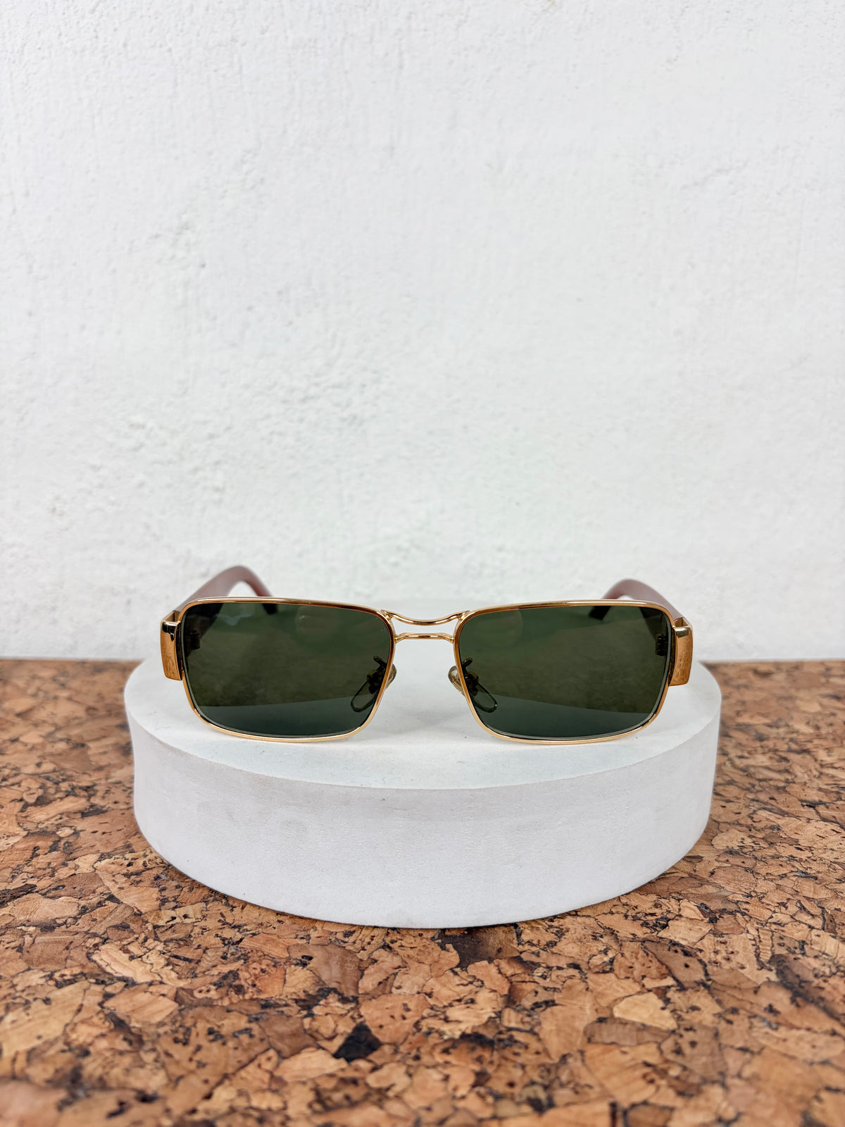 A pair of square sunglasses with gold metal frames and dark green lenses, resting on a round white display pedestal over a cork surface against a white textured wall.