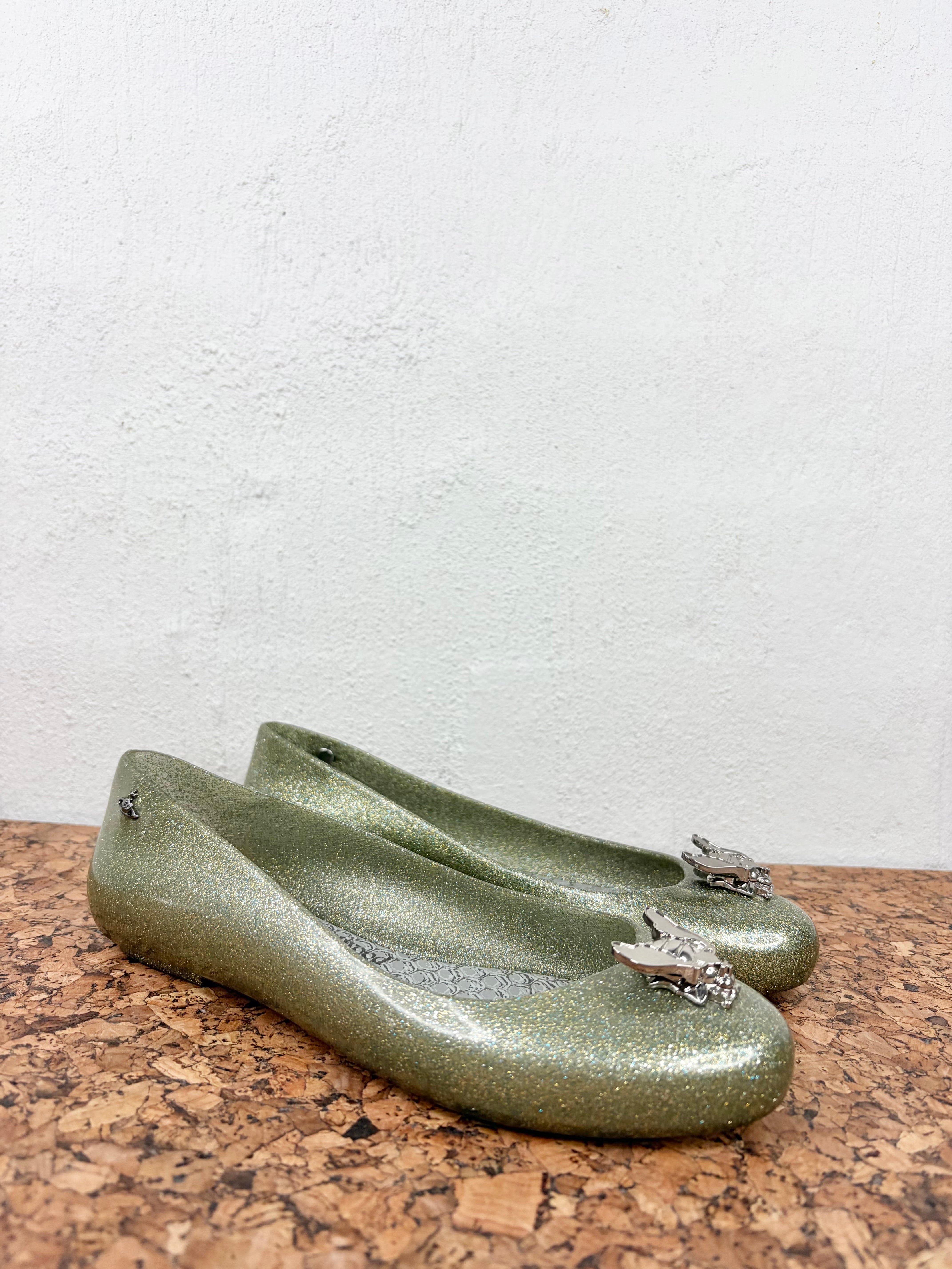 A pair of olive green glittery ballet flats with silver horse-shaped embellishments on the toes, resting on a cork surface against a white textured wall.