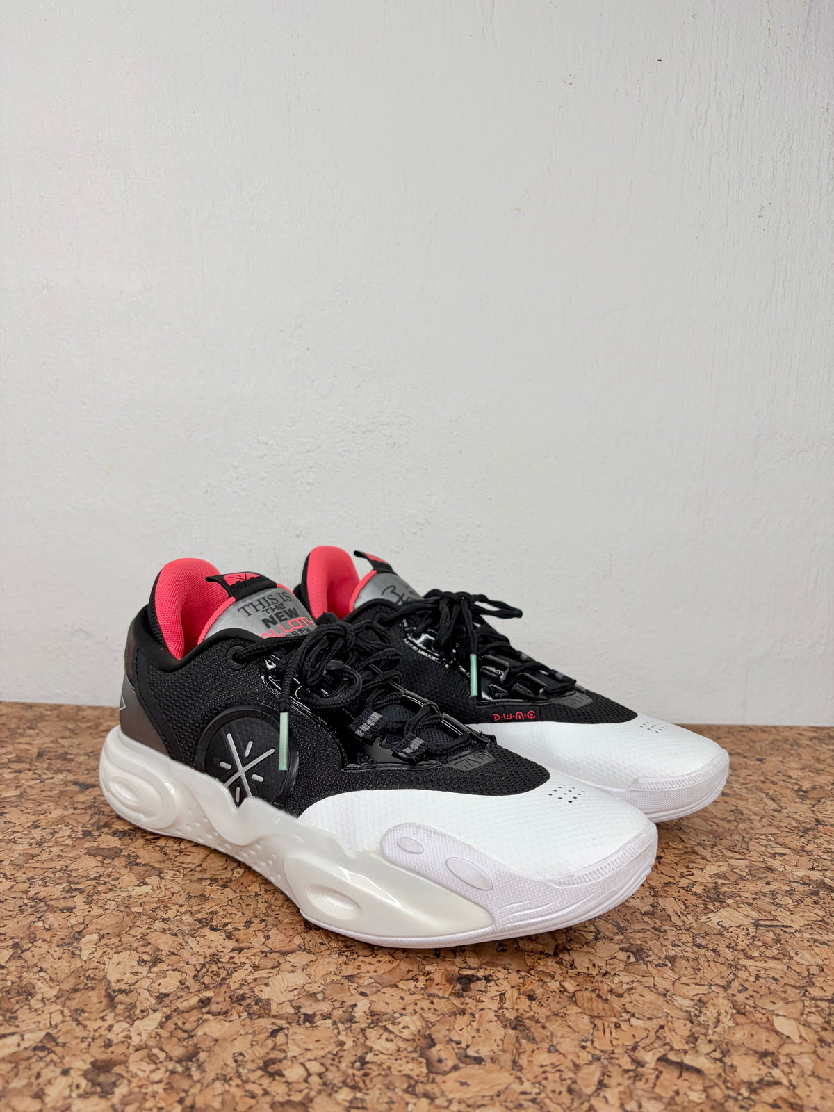 A pair of black and white athletic sneakers with bright pink accents on the tongue lining and a small green accent near the lace area, displayed on a brown cork surface against a white wall.