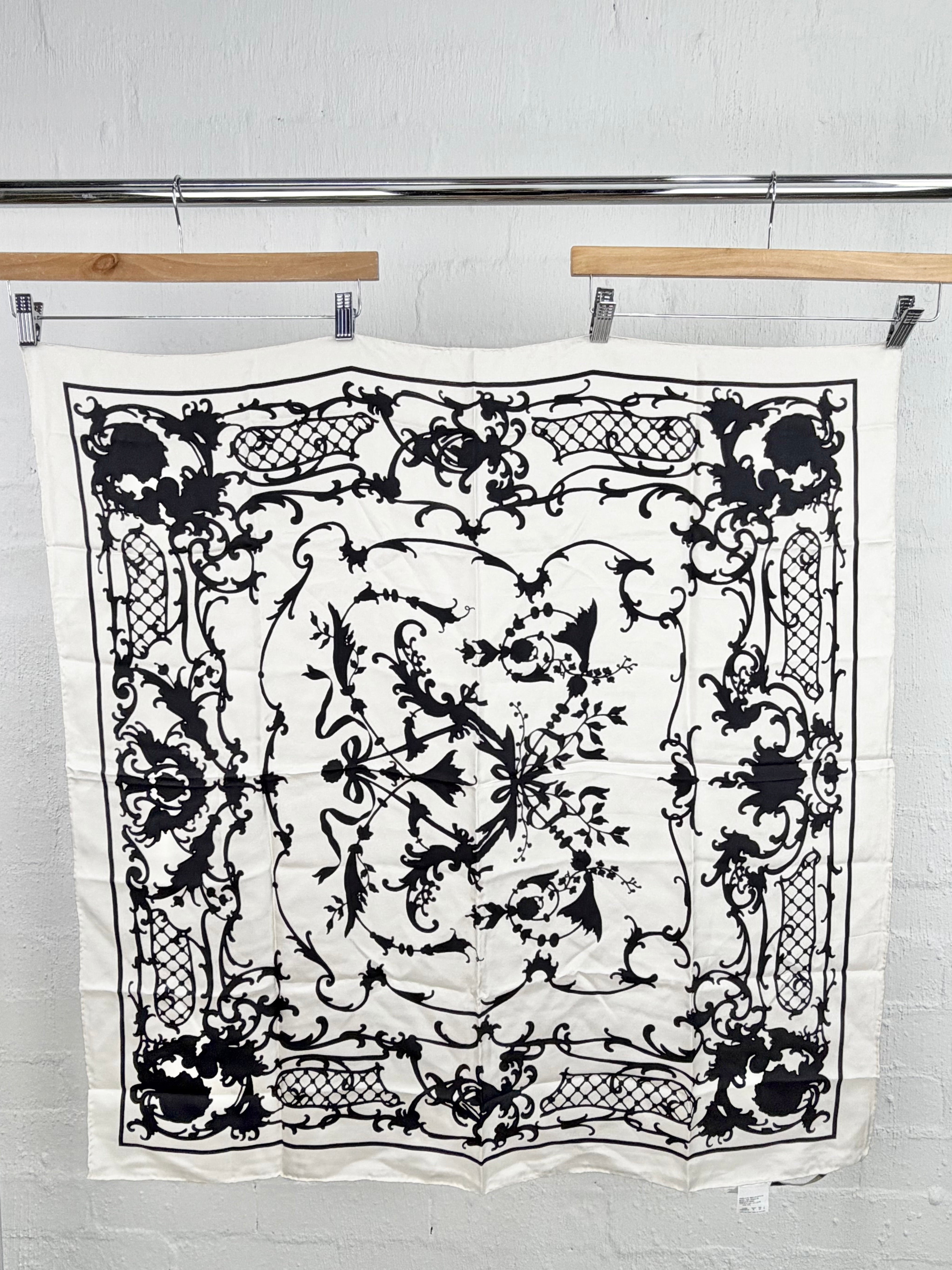 A square white scarf with an intricate black baroque-style floral and scroll pattern, hanging from wooden hangers on a metal rack against a white brick wall.