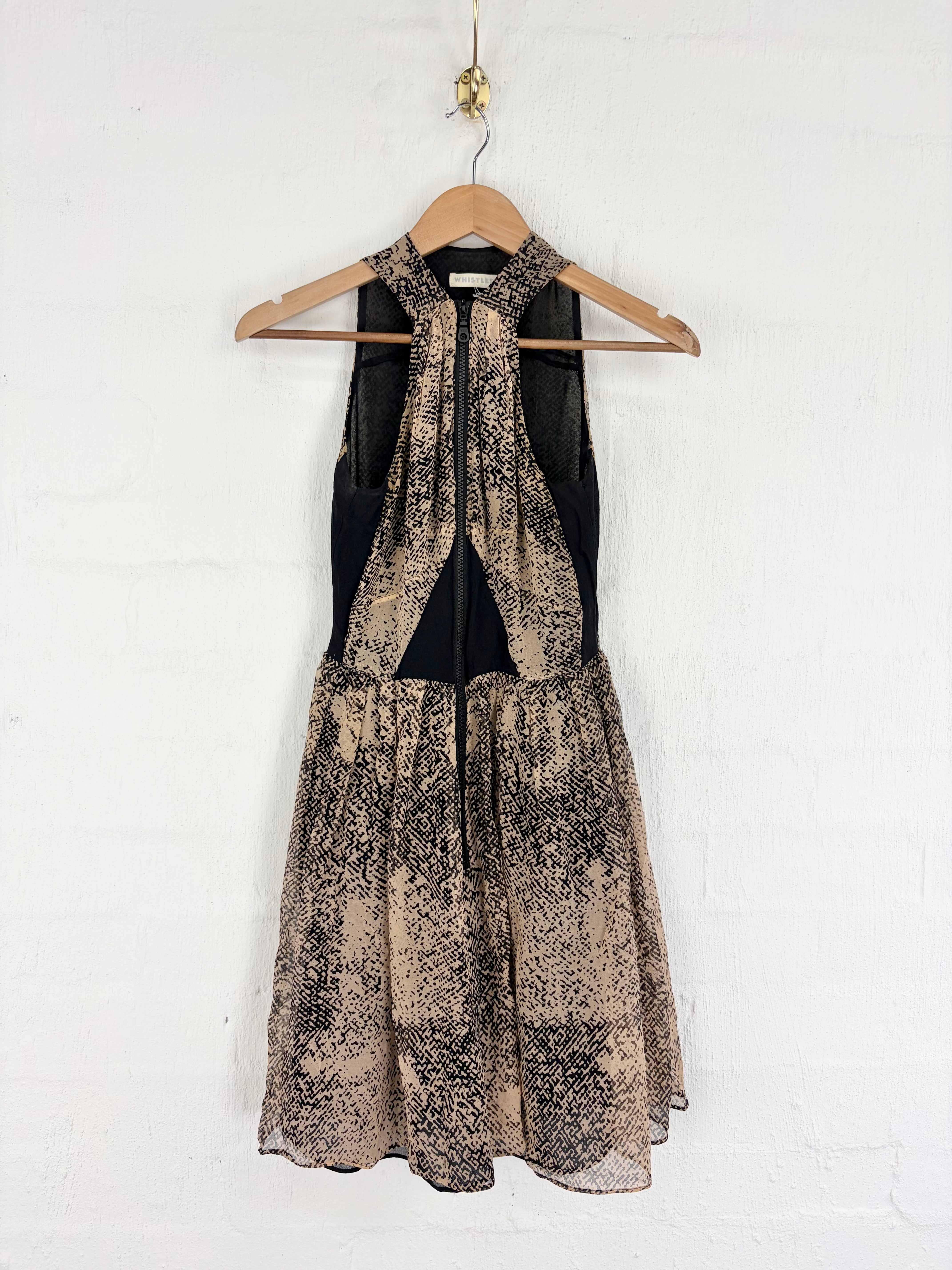 A sleeveless midi dress with a black and beige abstract patterned skirt and sheer black mesh overlay, hanging on a wooden hanger against a white painted brick wall.