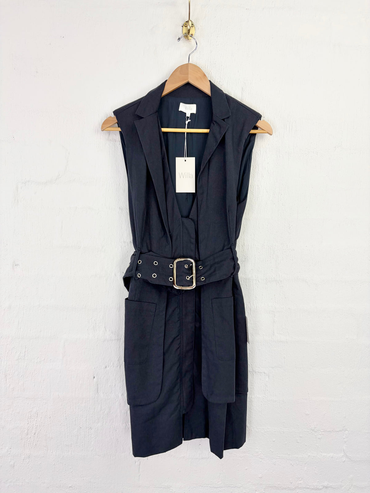 A sleeveless black belted vest dress with notched lapels and front patch pockets, hanging on a wooden hanger against a white painted brick wall.