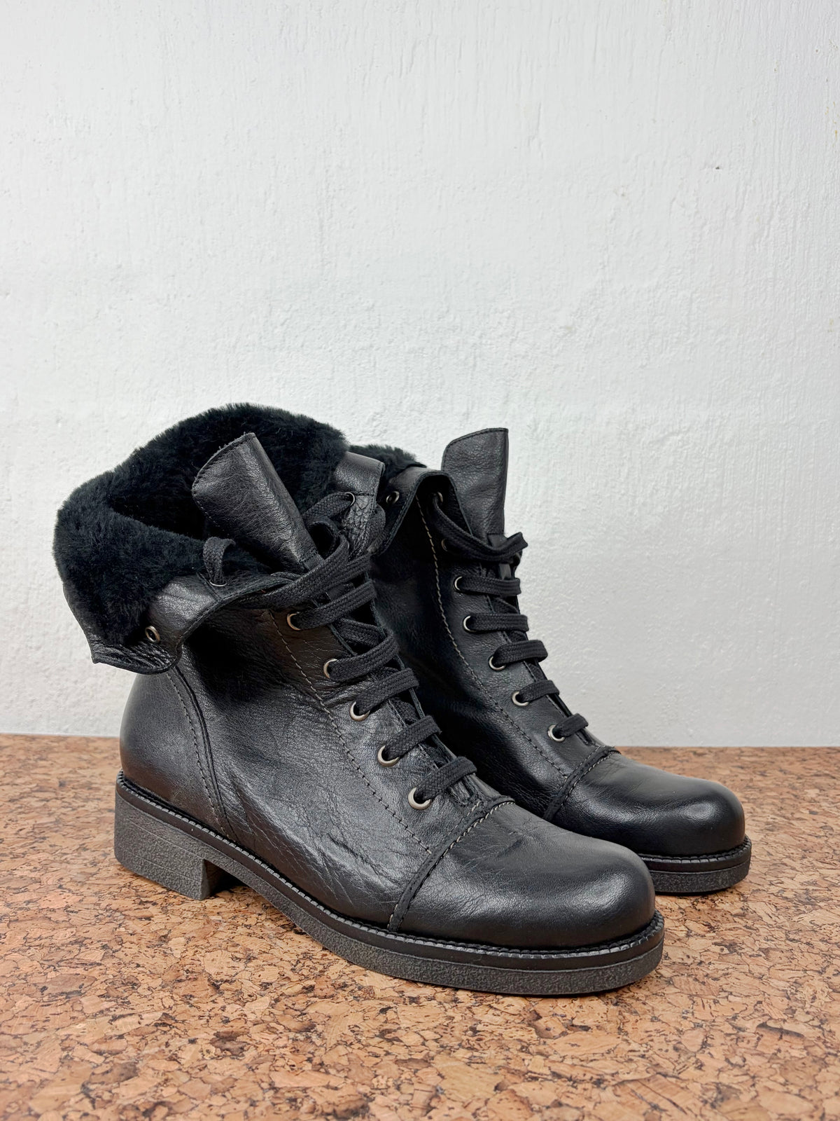 A pair of black leather lace-up ankle boots with a plush black shearling-style collar, resting on a brown textured surface against a white wall.