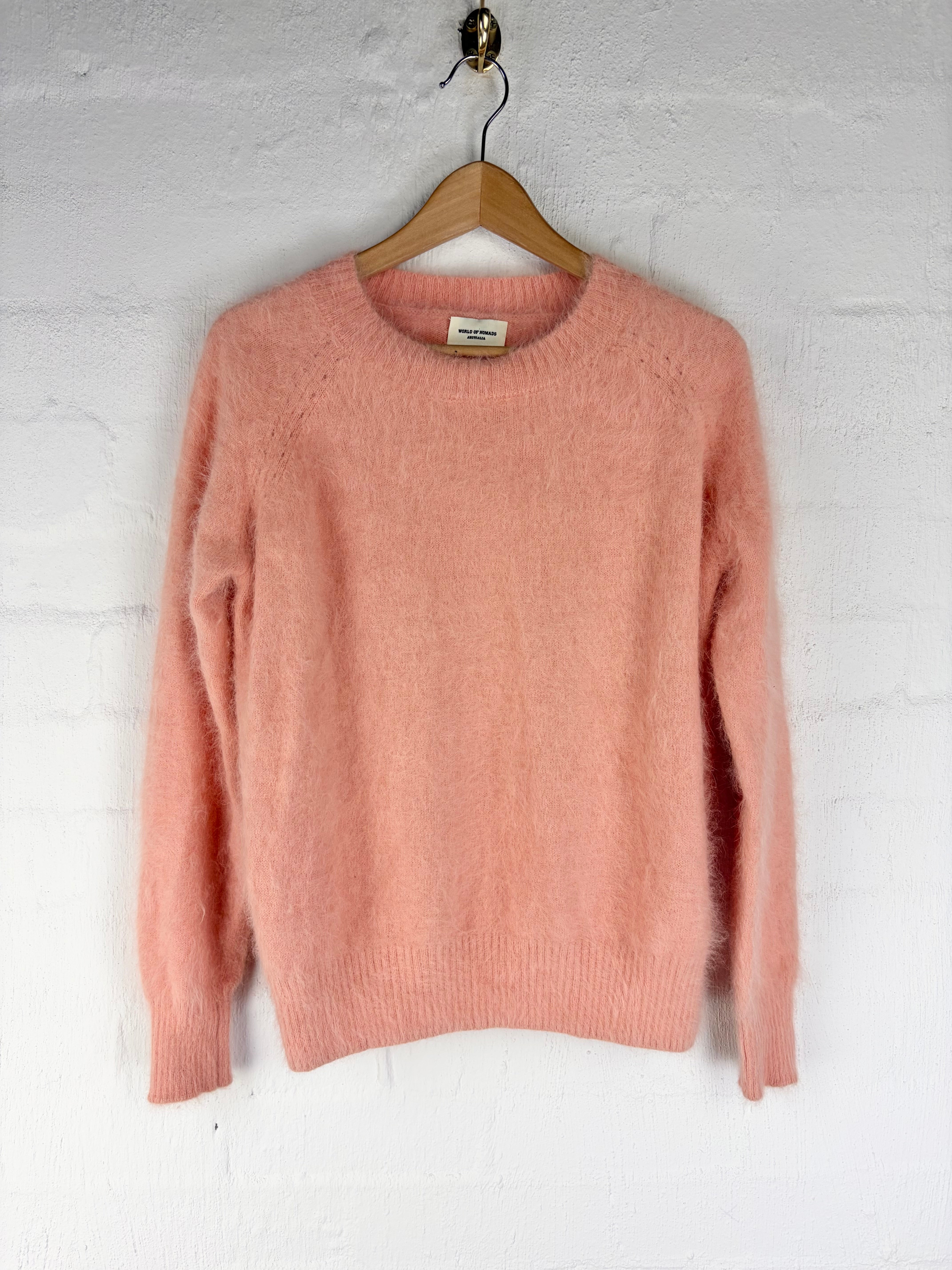 A dusty pink knit crewneck sweater hanging on a wooden hanger against a white painted brick wall.