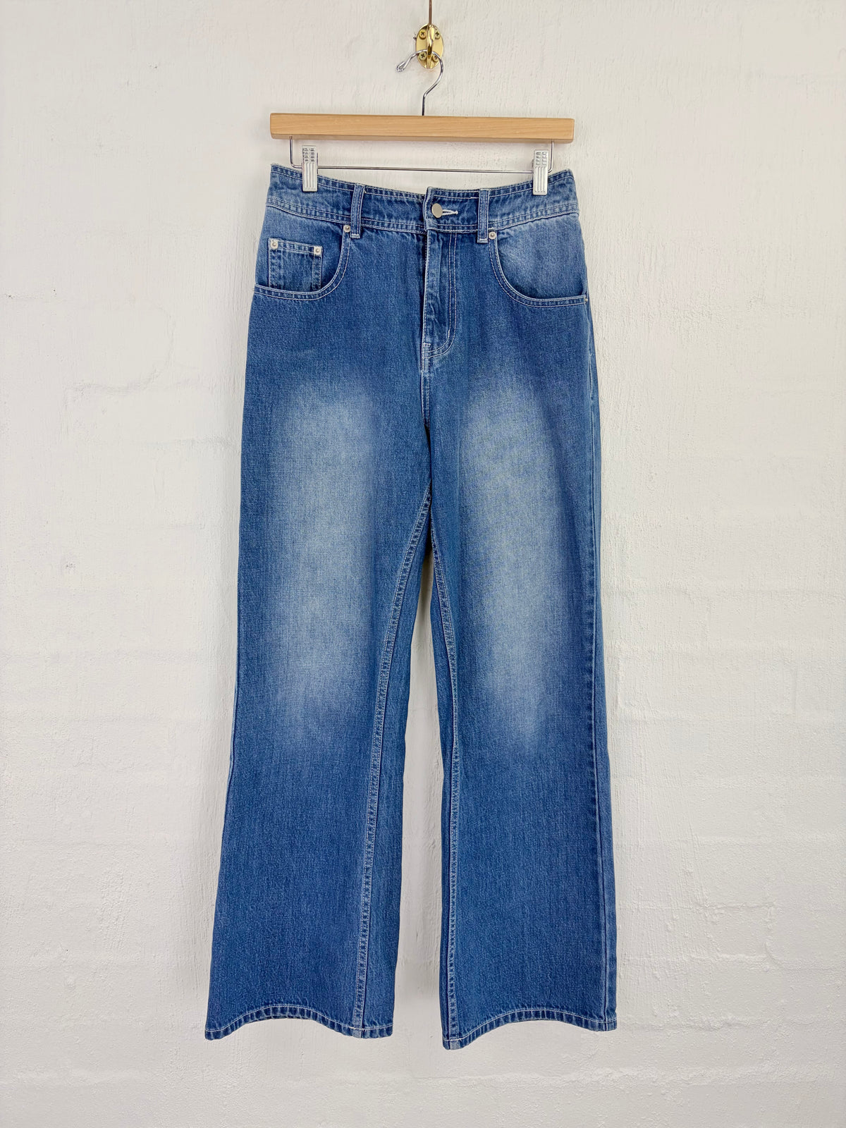 Medium wash blue denim flare jeans hanging on a wooden hanger against a white painted brick wall.