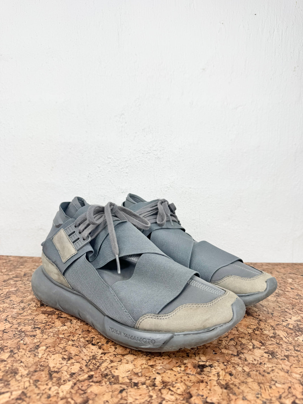 A pair of gray wrap-around lace-up sneakers with beige toe and heel accents, resting on a brown speckled stone surface against a white wall.