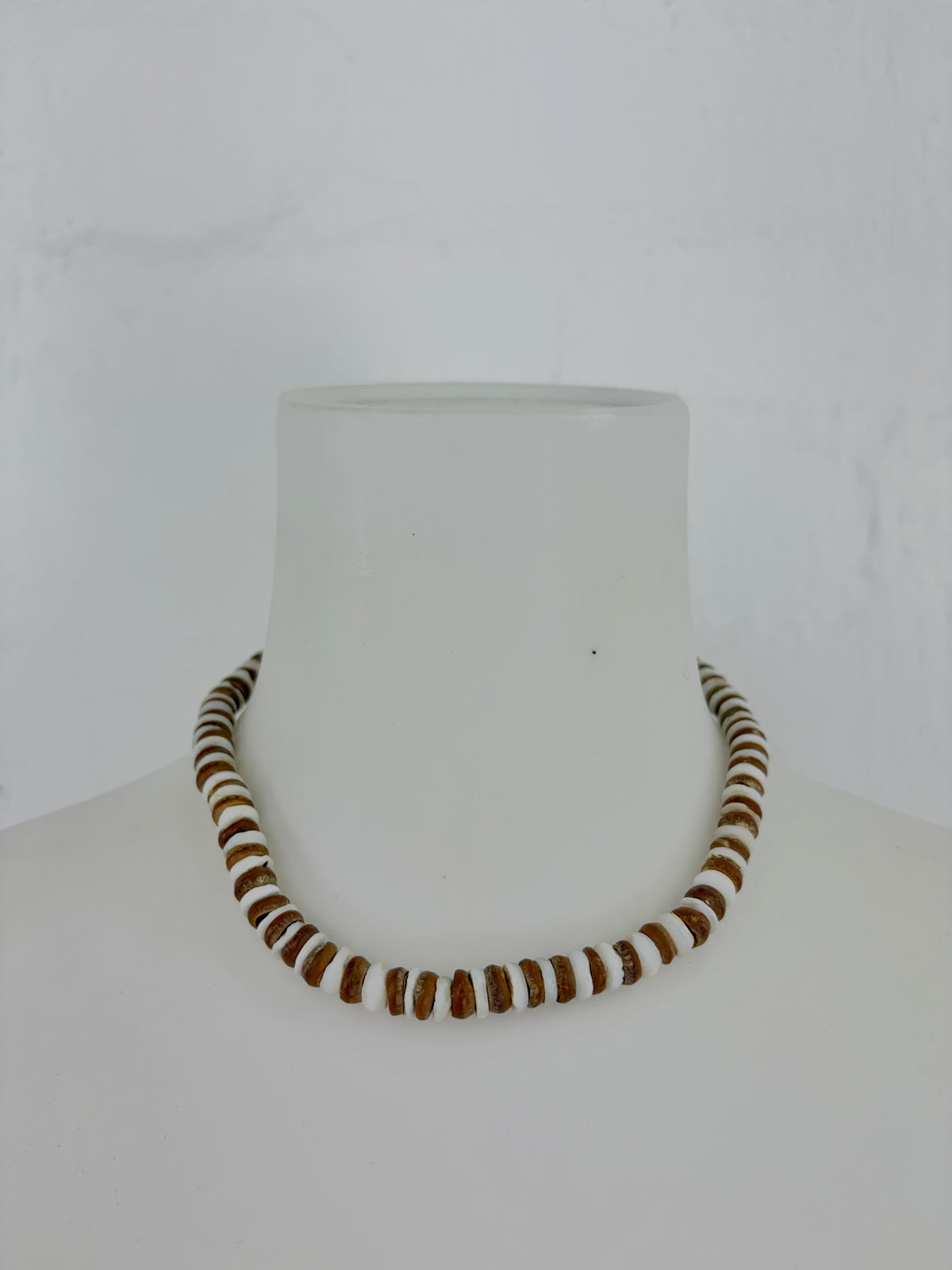 A beaded necklace made of alternating brown and white cylindrical beads, displayed on a white mannequin bust against a white background.