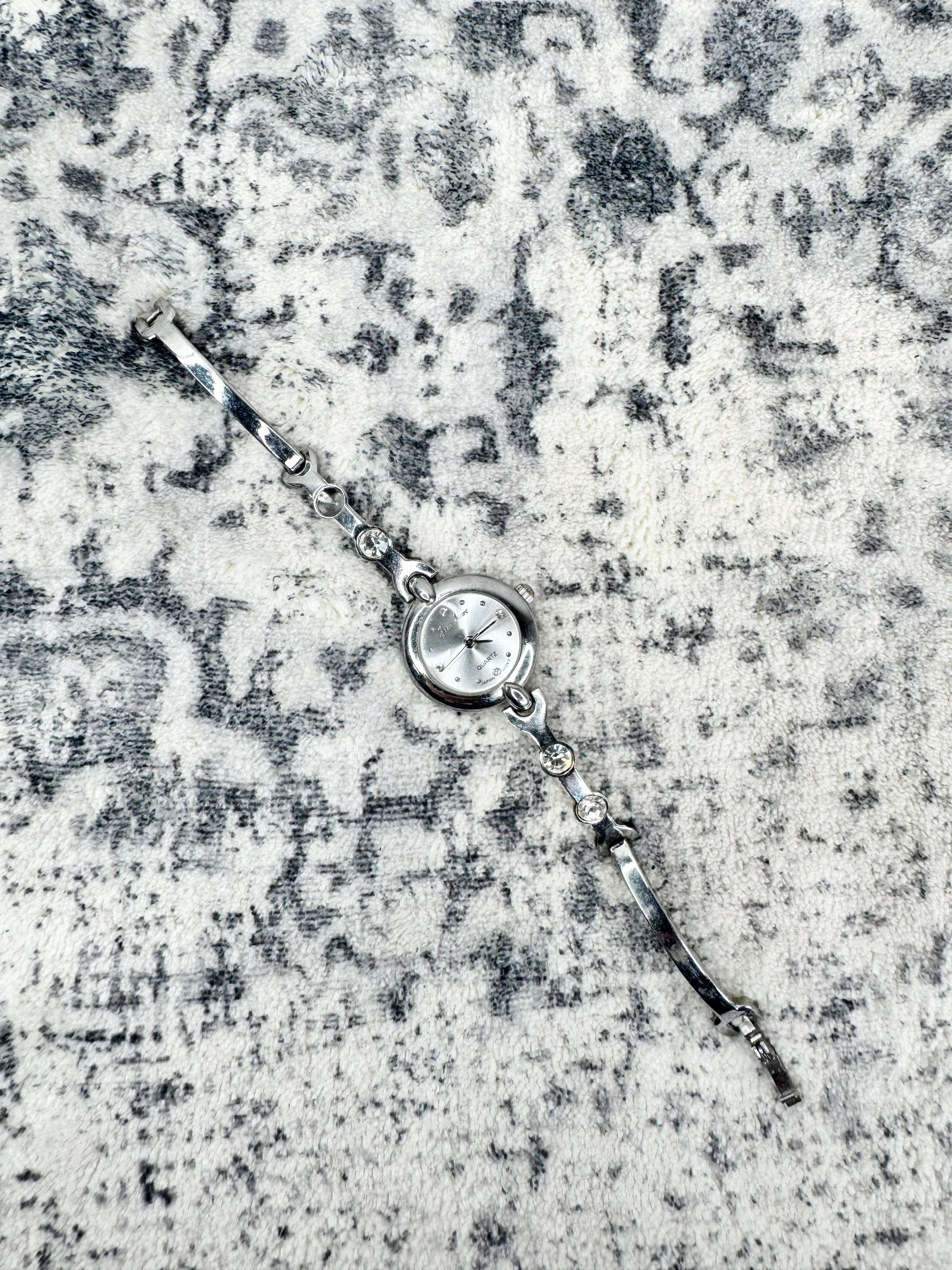 A silver-toned watch with a round bezel and clear crystal face, laid diagonally on a textured gray and white patterned fabric background.