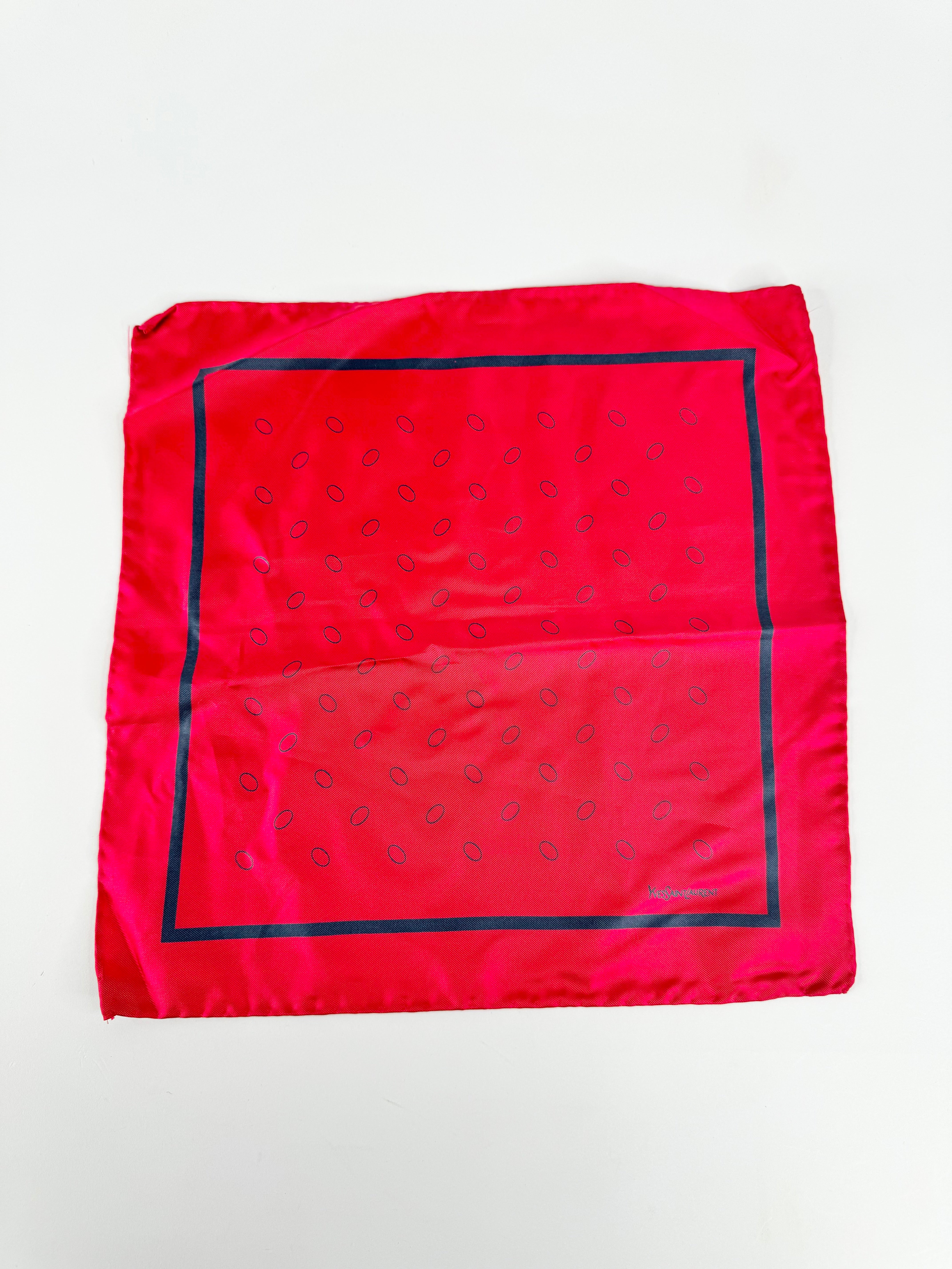 A folded red silk bandana featuring a navy blue border and a repeating paisley pattern, laid flat on a white background.