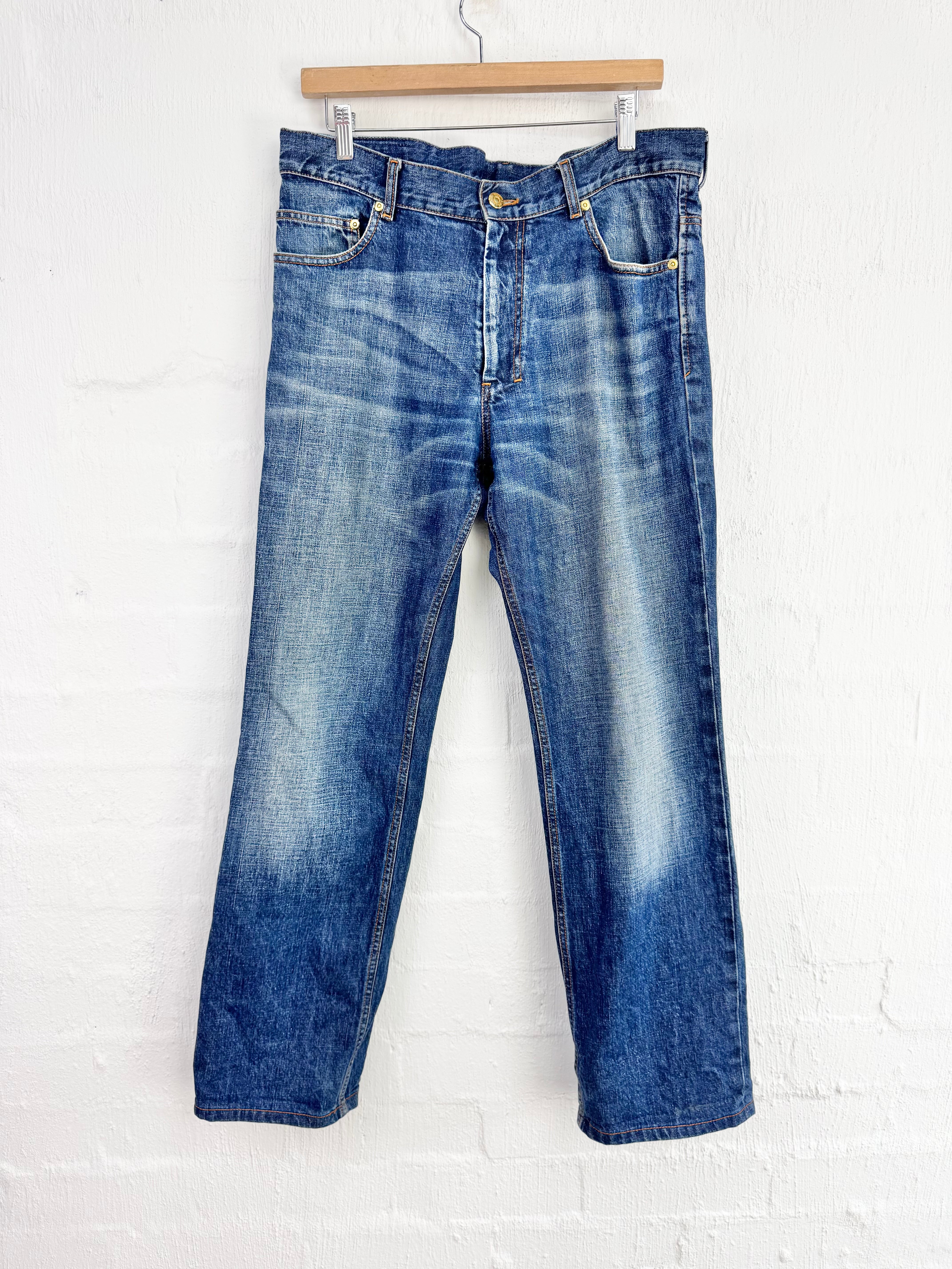 A pair of blue denim jeans with a faded wash, hanging from a wooden hanger against a white painted brick wall.