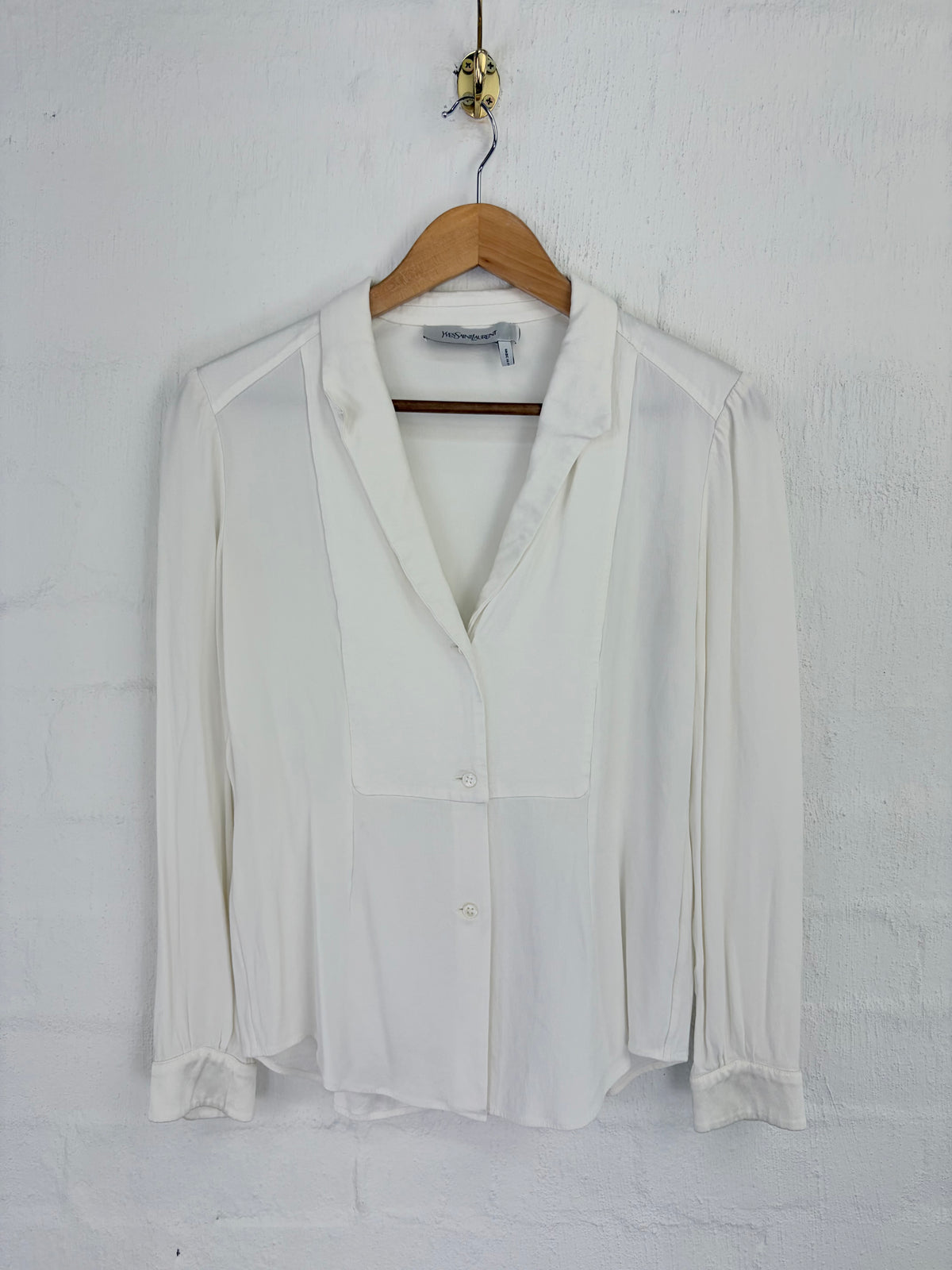 A white long-sleeve blouse with a notched lapel and button front, hanging on a wooden hanger against a white painted brick wall.