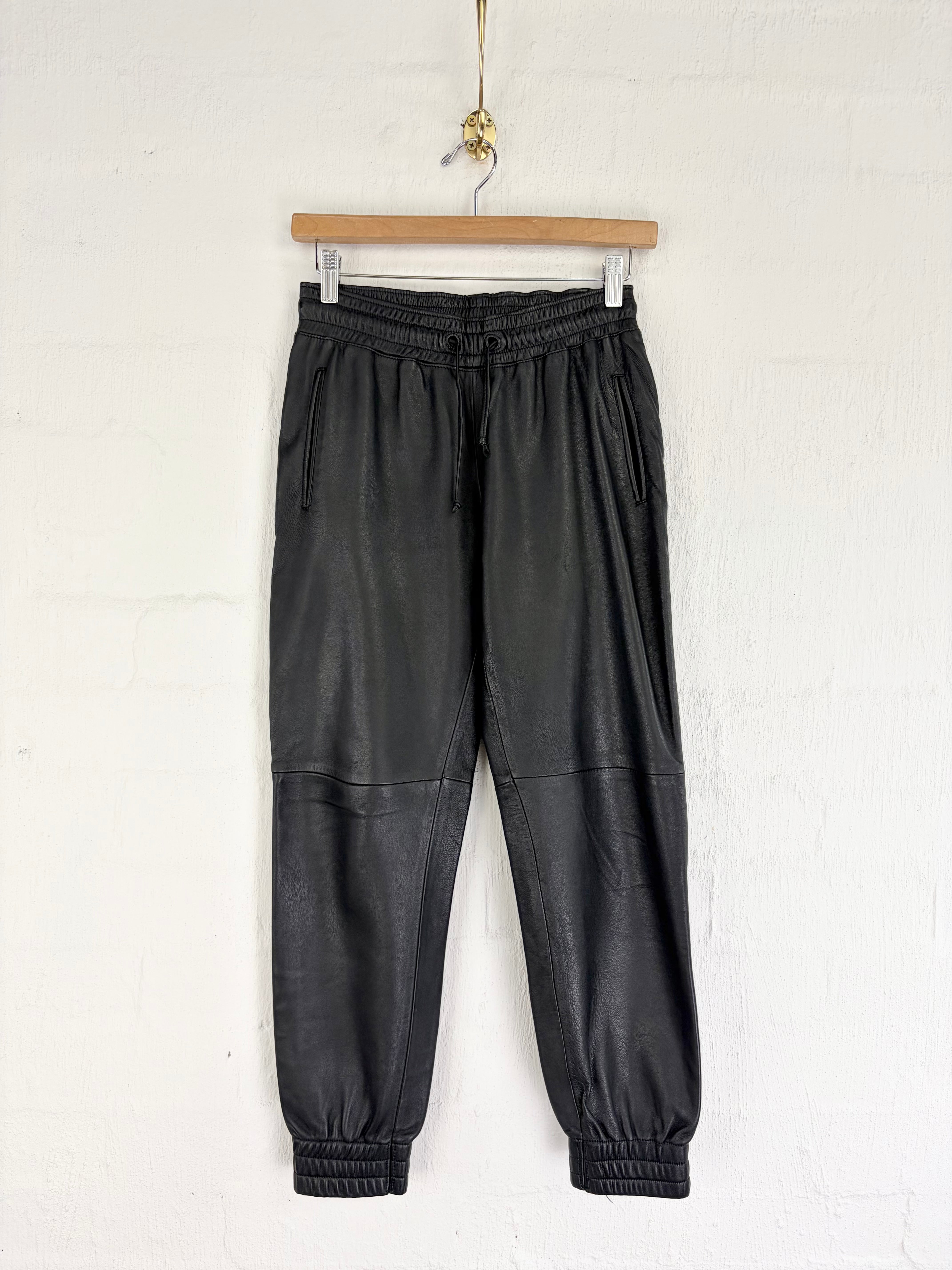 Black faux leather jogger pants hanging from a wooden hanger against a white brick wall.