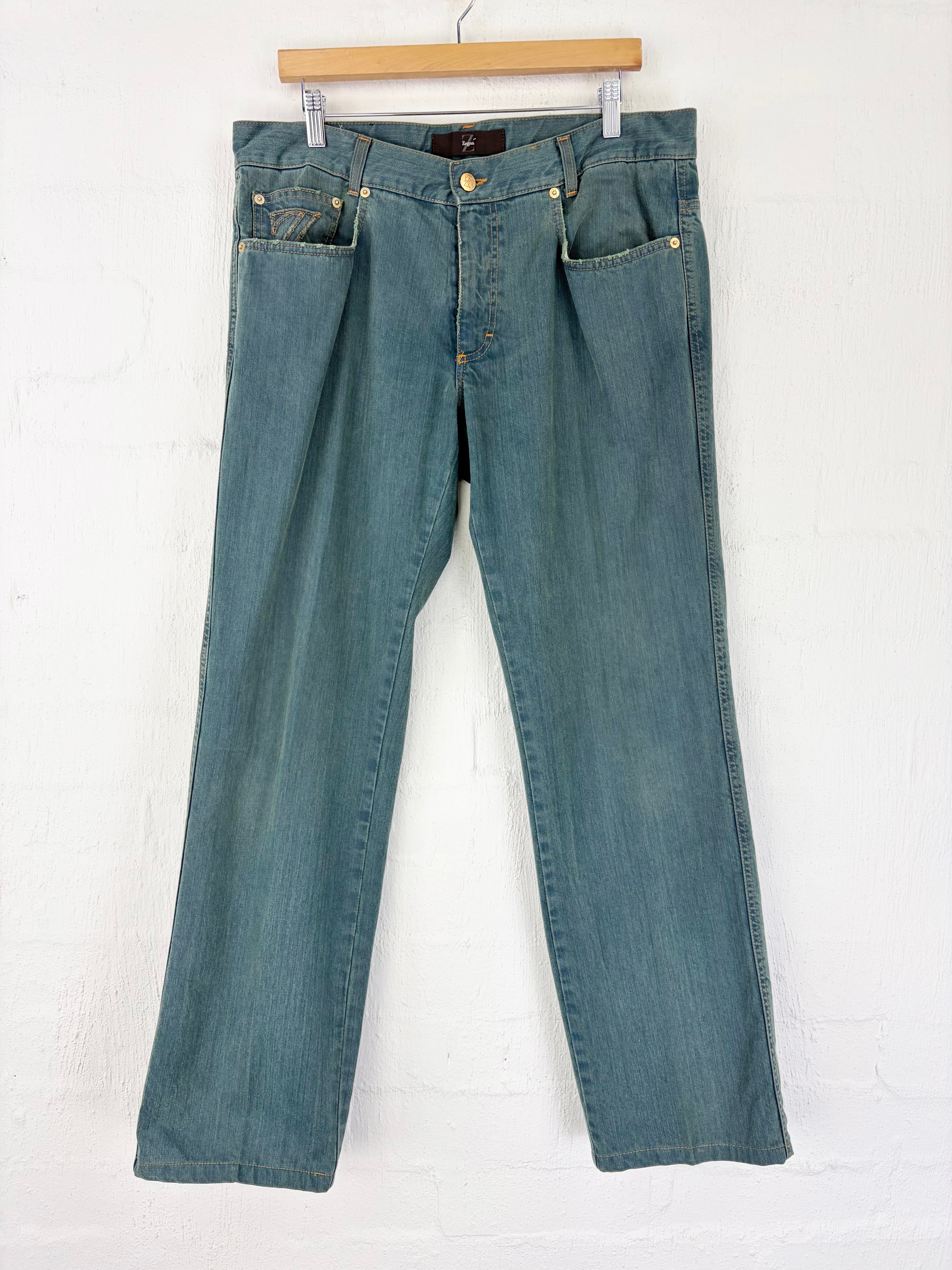 A pair of blue denim jeans hanging from a wooden hanger against a white painted brick wall.