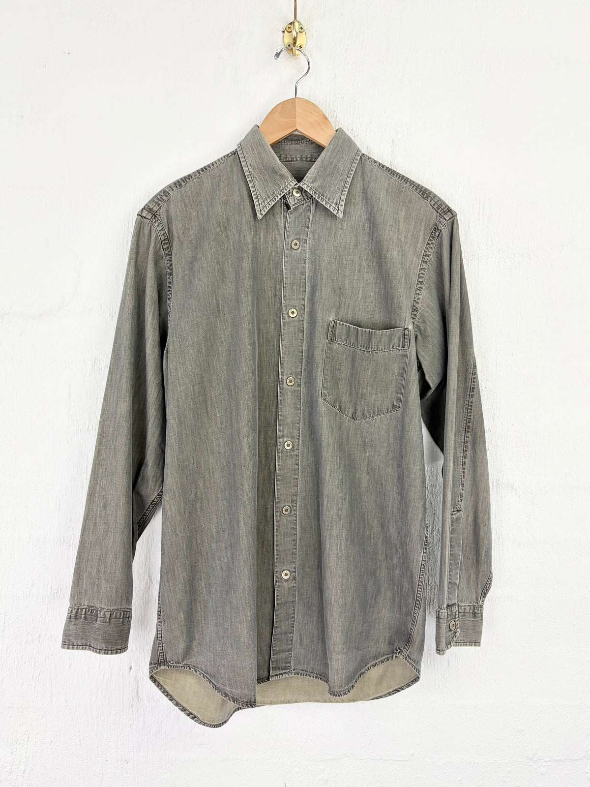 A gray washed denim long-sleeve button-up shirt hanging on a wooden hanger against a white textured wall.