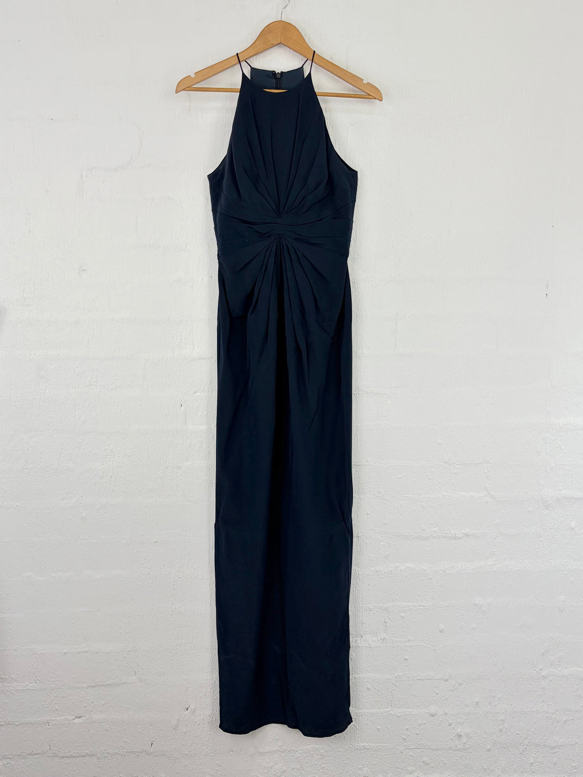 A sleeveless navy blue halter maxi dress with a twisted front detail, hanging on a wooden hanger against a white painted brick wall.
