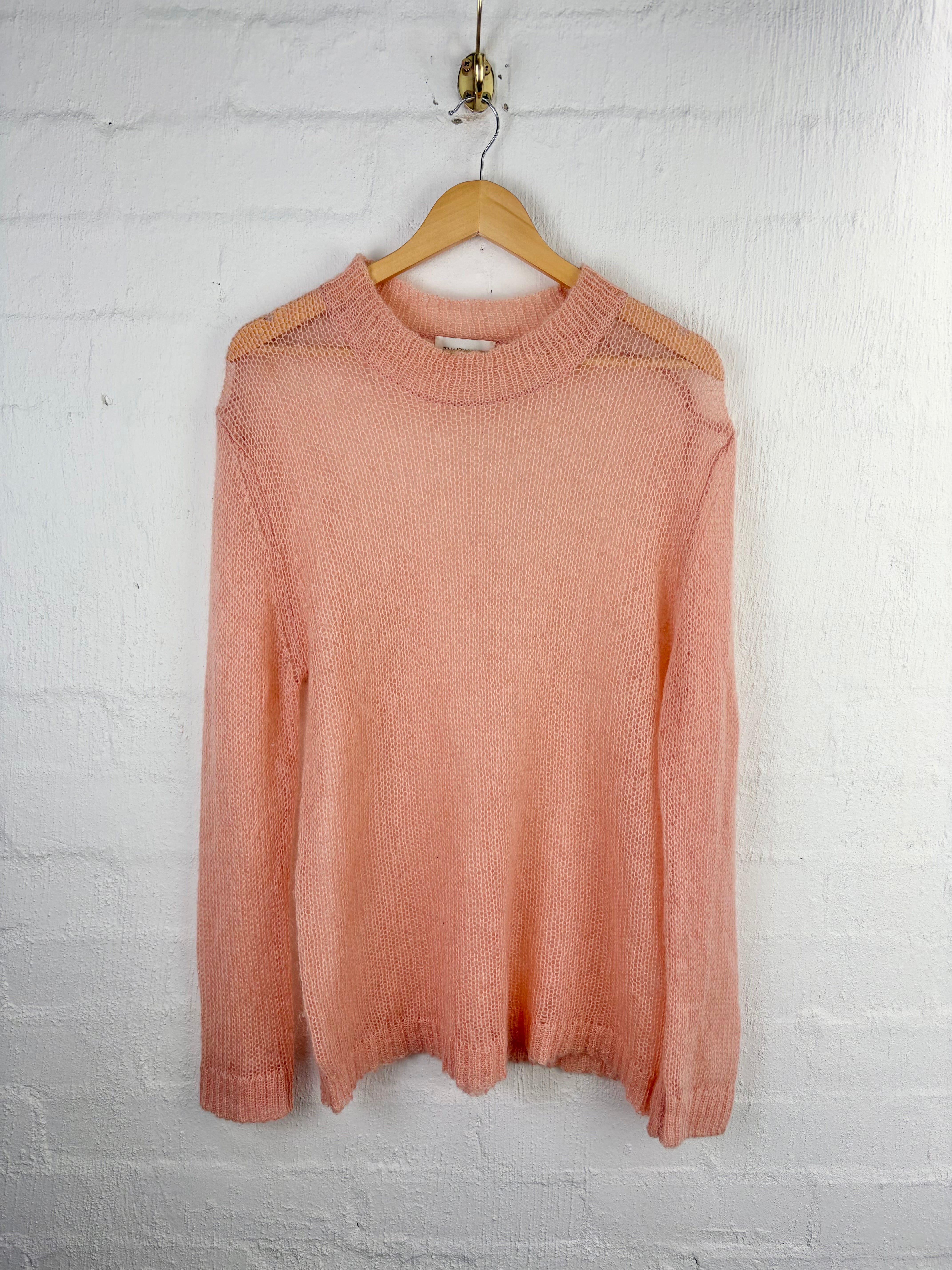 A peach-colored knit sweater with shoulder cutouts, hanging on a wooden hanger against a white painted brick wall.
